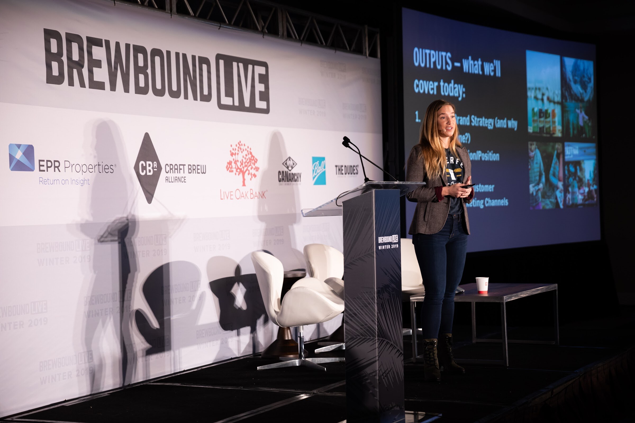 Woman at a lectern on a stage with the Brewbound Live backdrop and a projected slide headed Outputs.