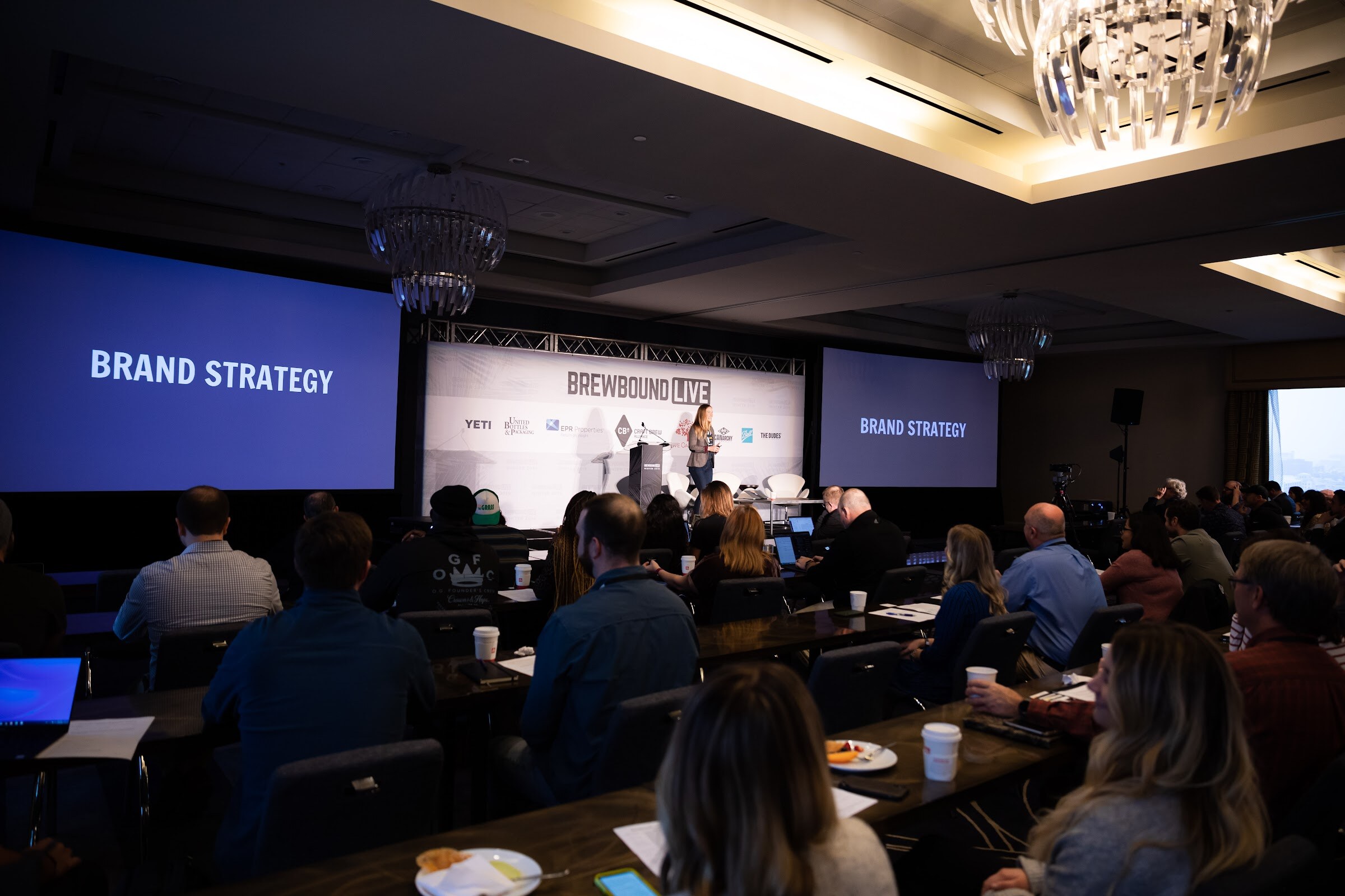 Wide view of a darkened ballroom with seated attendees and two screens reading Brand Strategy.