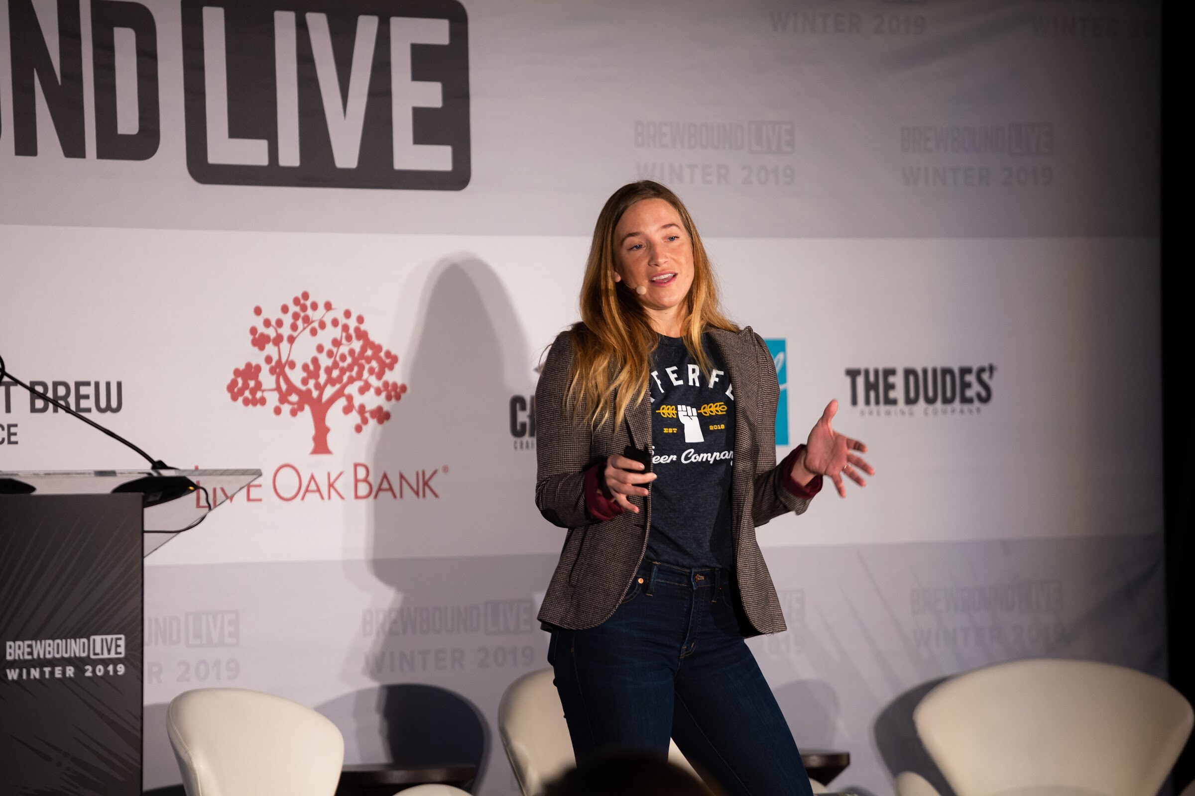 Woman in a tweed blazer and dark T-shirt gesturing on stage in front of the Brewbound Live backdrop.