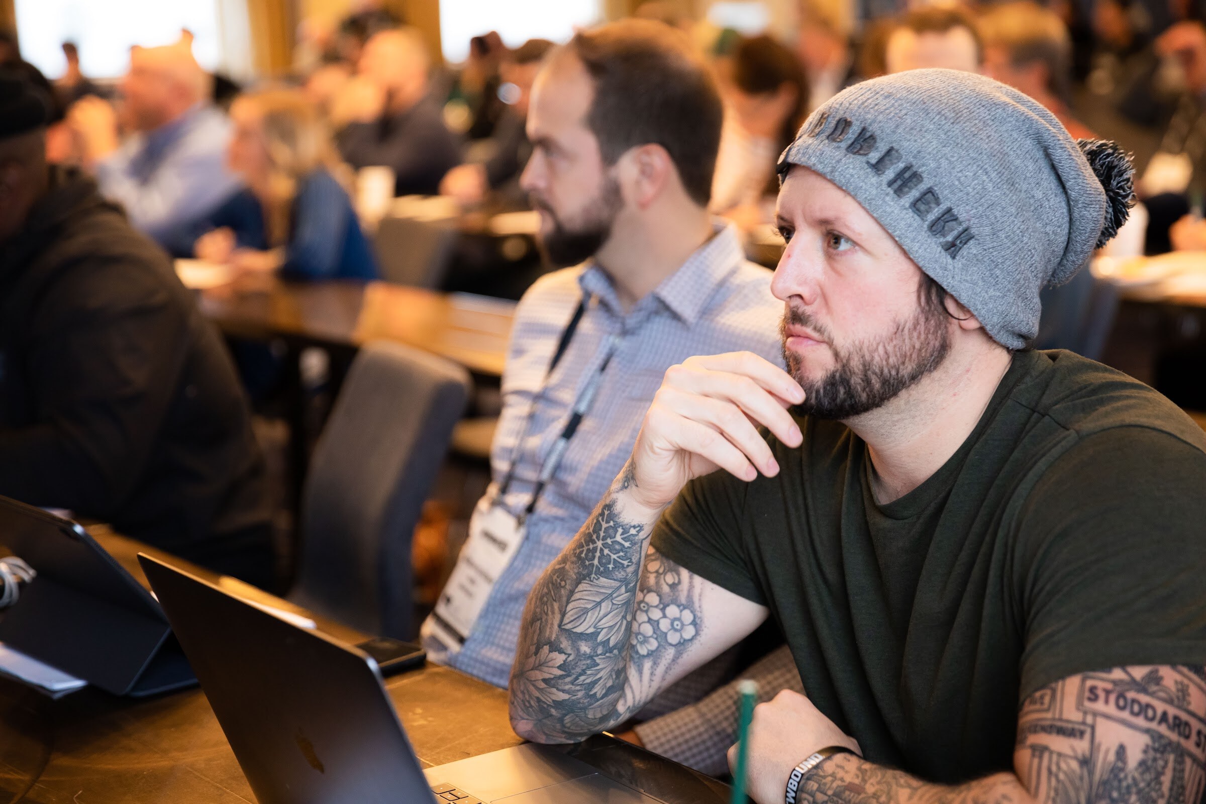 Tattooed man in a gray beanie sitting at a table with a laptop, looking toward the stage.