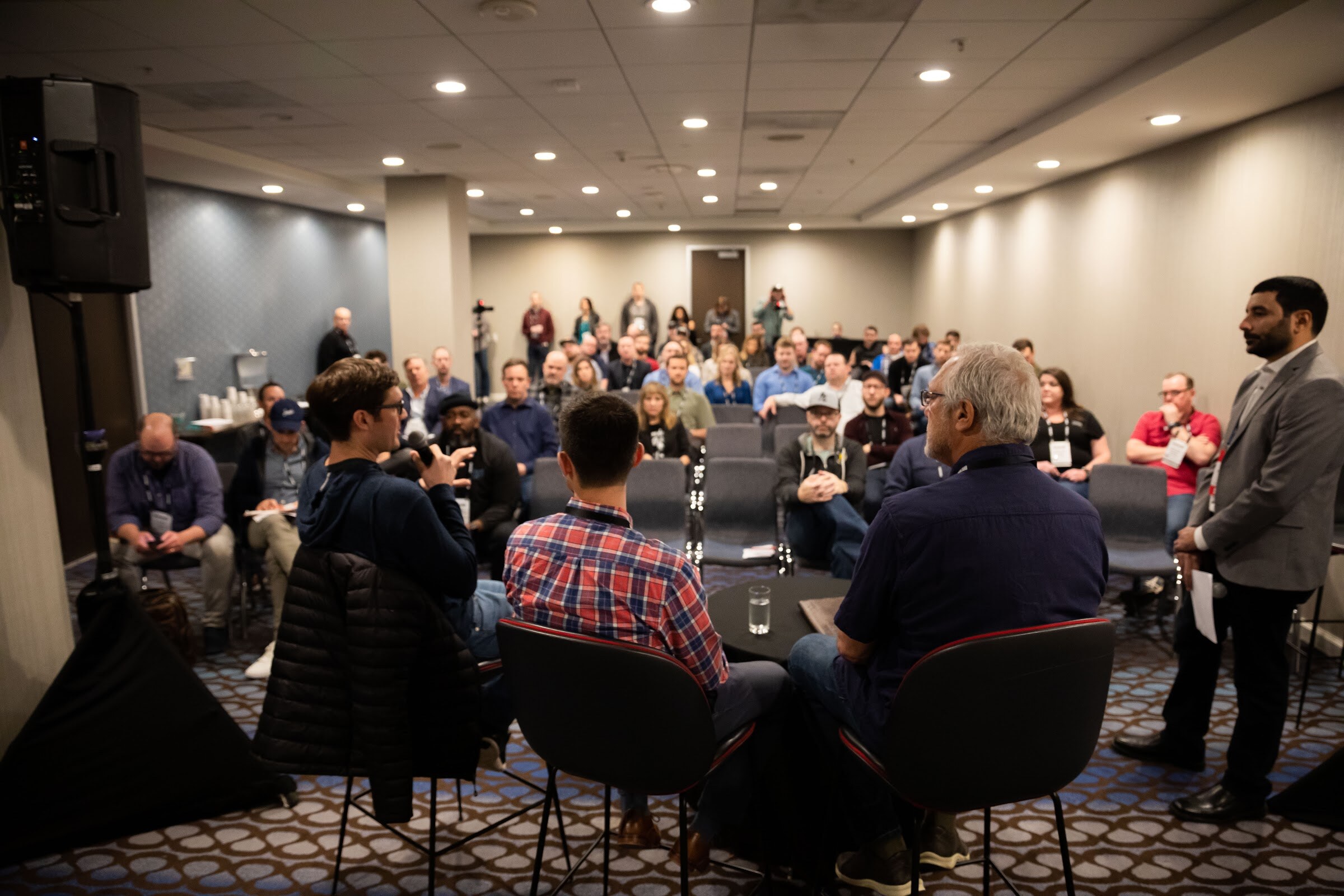 Rows of seated attendees facing a panel with a standing moderator in a small meeting room.