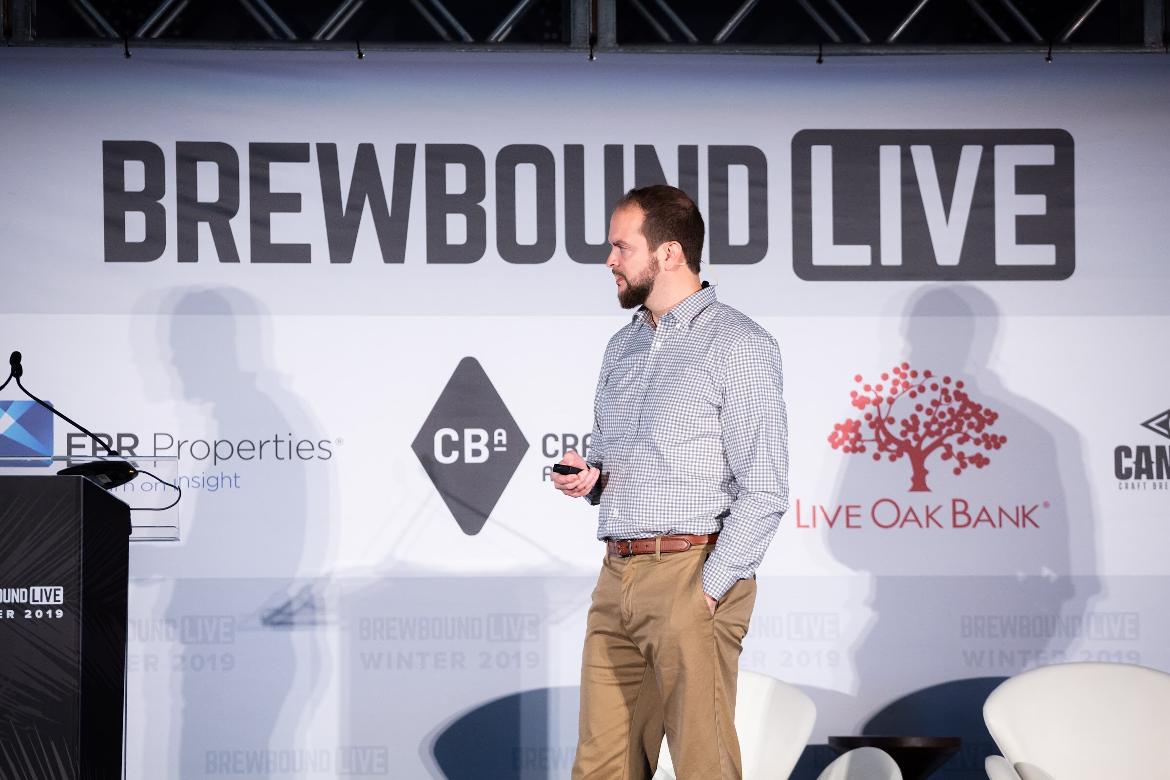 Man in a checked shirt and khakis standing on stage in front of the Brewbound Live backdrop.