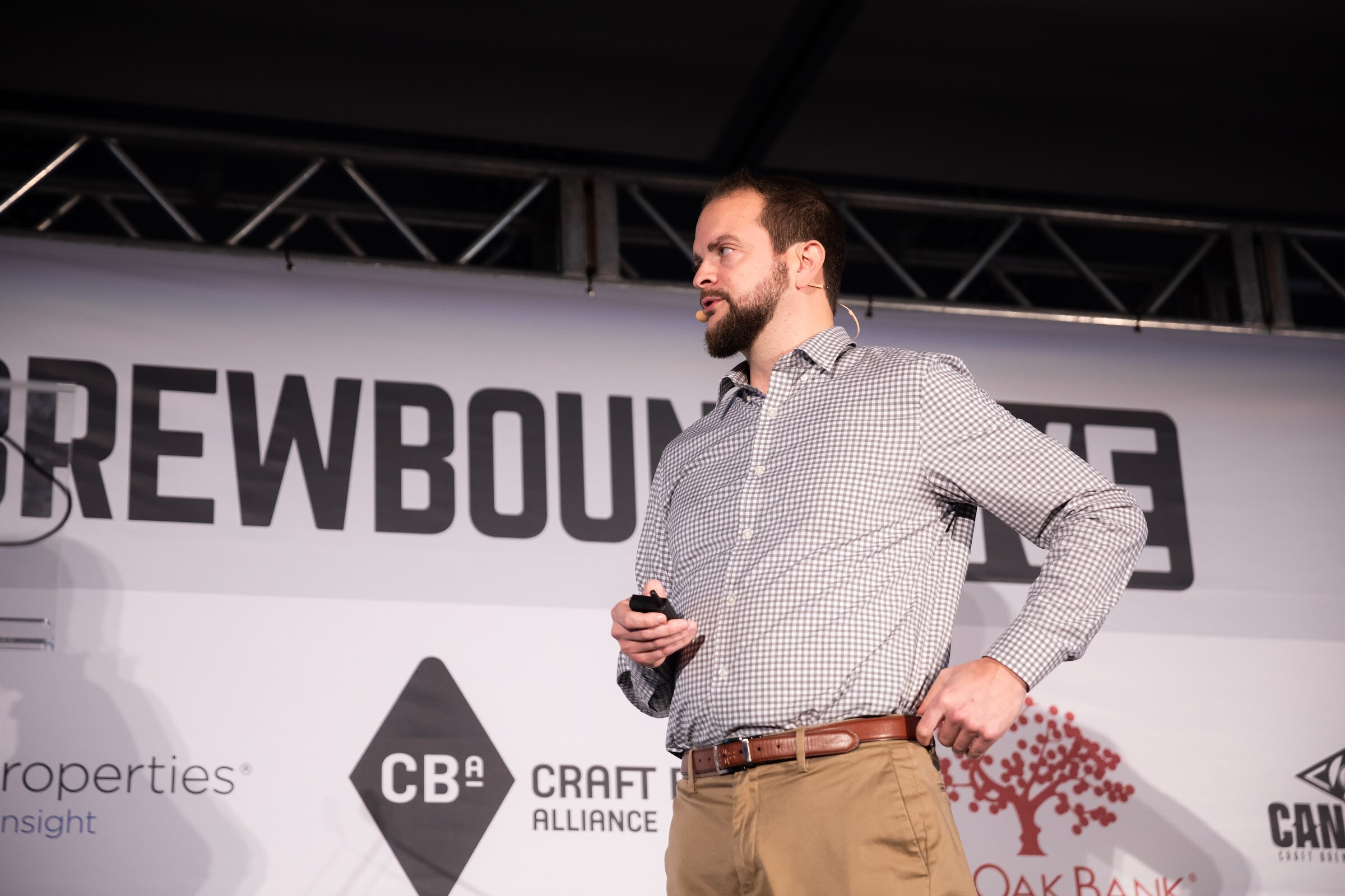 Man in a gray checked shirt standing at a lectern with one hand on his hip in front of the Brewbound Live backdrop.