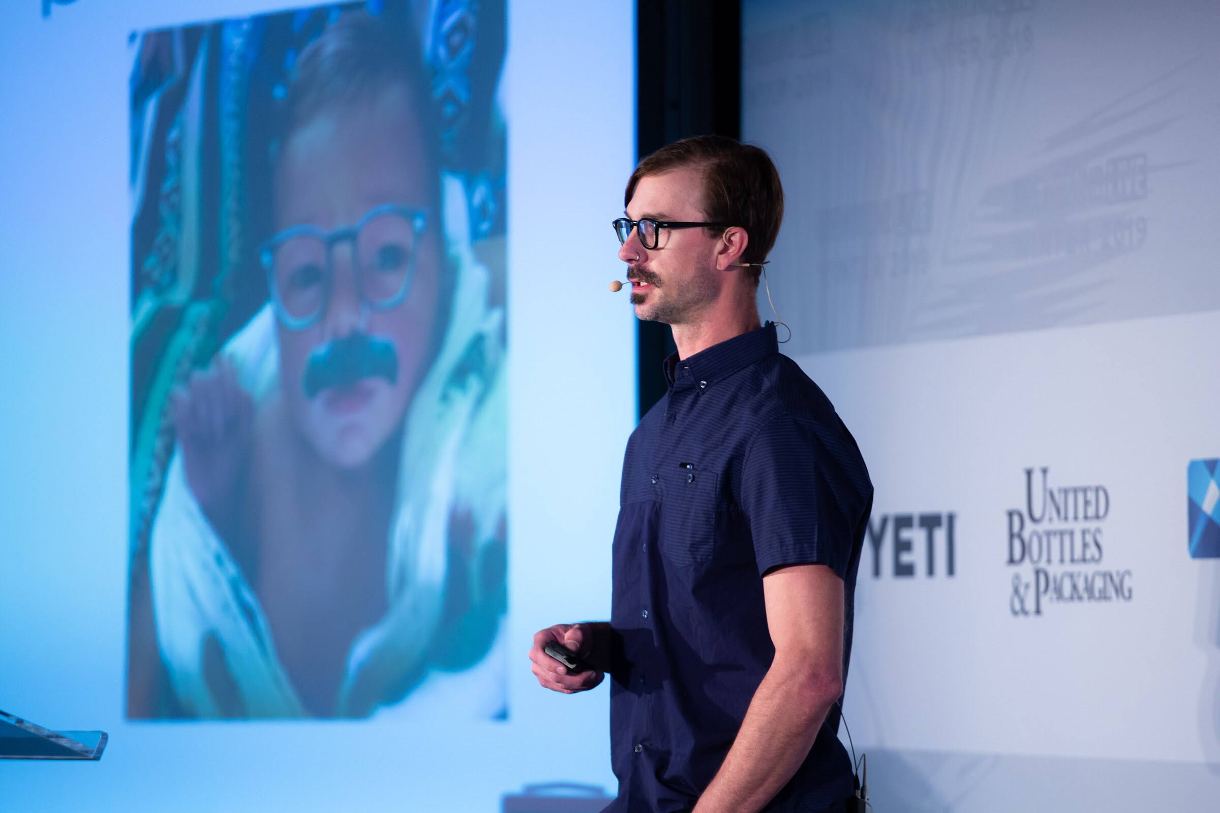 Man in glasses and a navy shirt on stage next to a large projected photo of a baby wearing glasses and a mustache sticker.