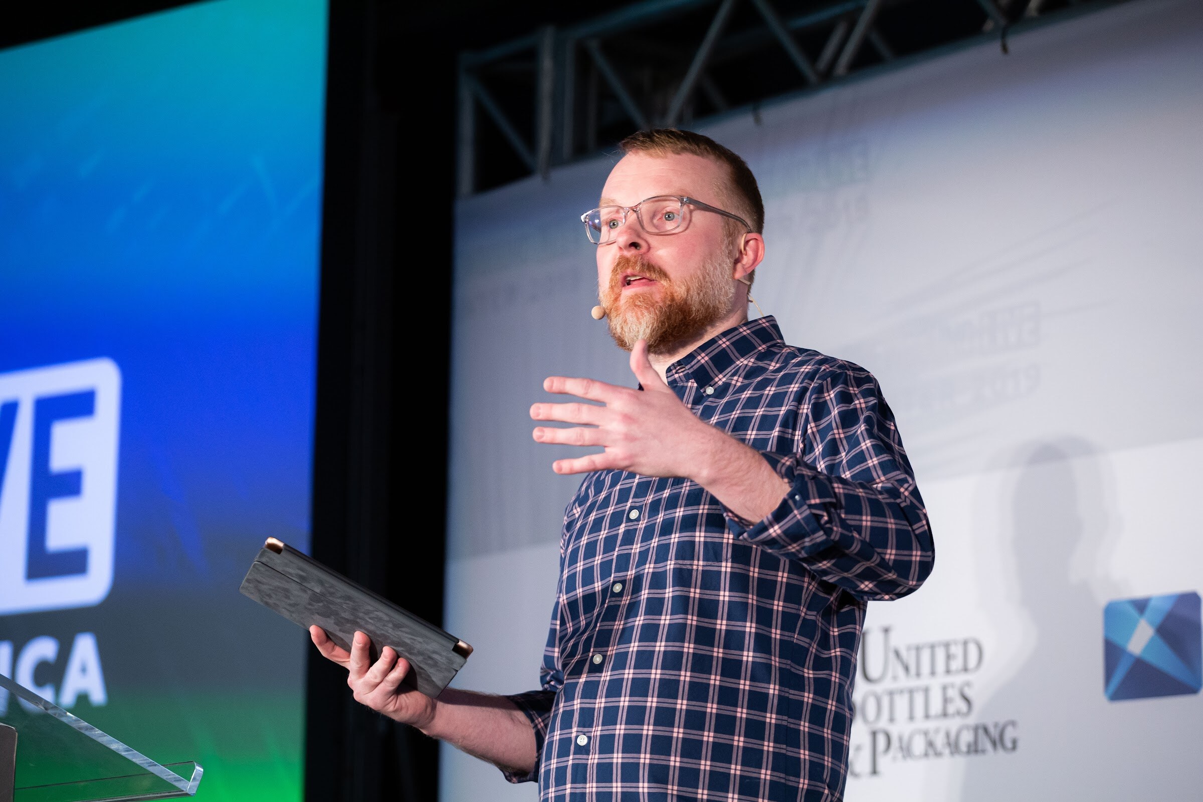 Bearded man in glasses and a plaid shirt holding a tablet and gesturing on stage.