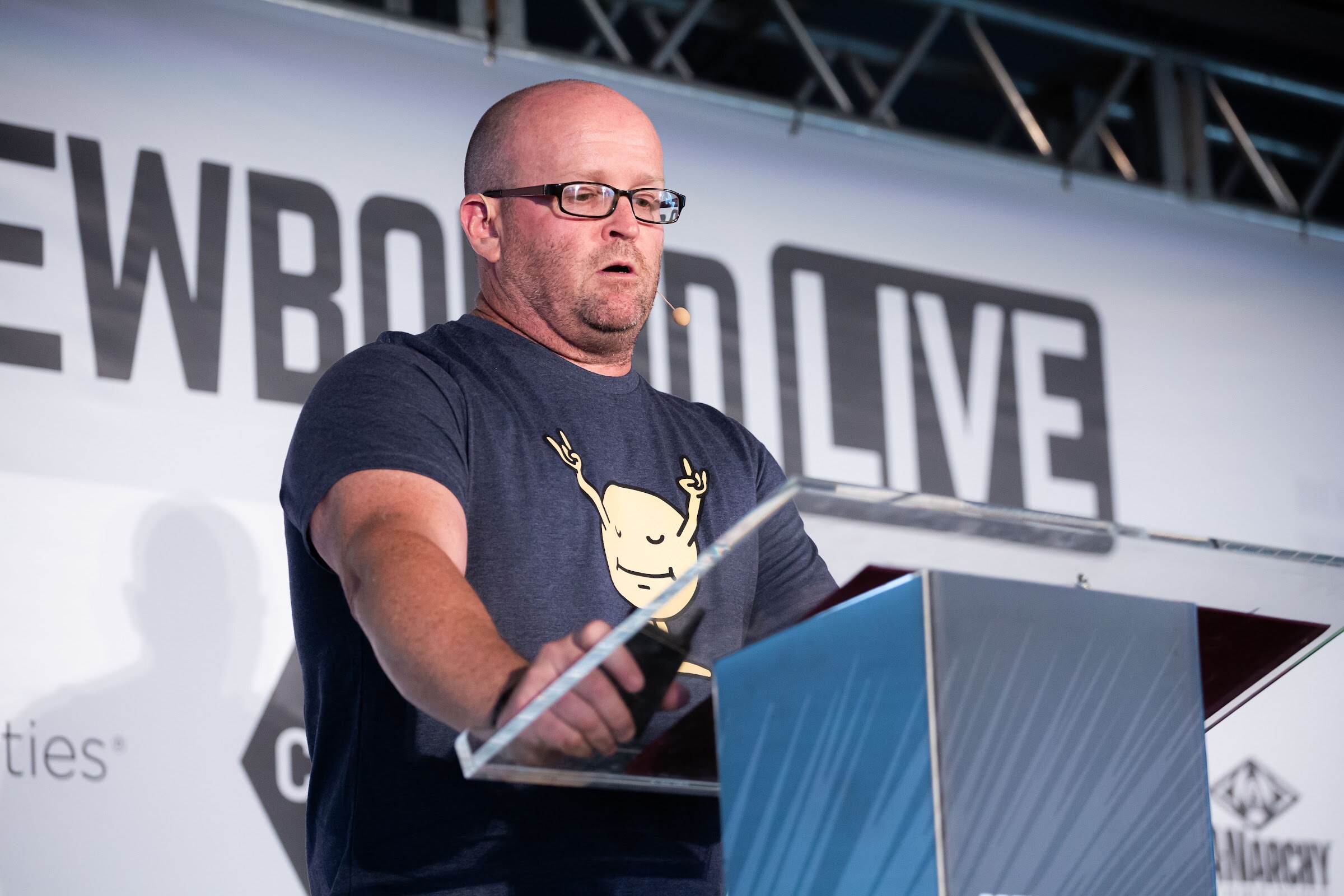 Bald man with glasses in a navy T-shirt speaking at a lectern in front of the Brewbound Live backdrop.