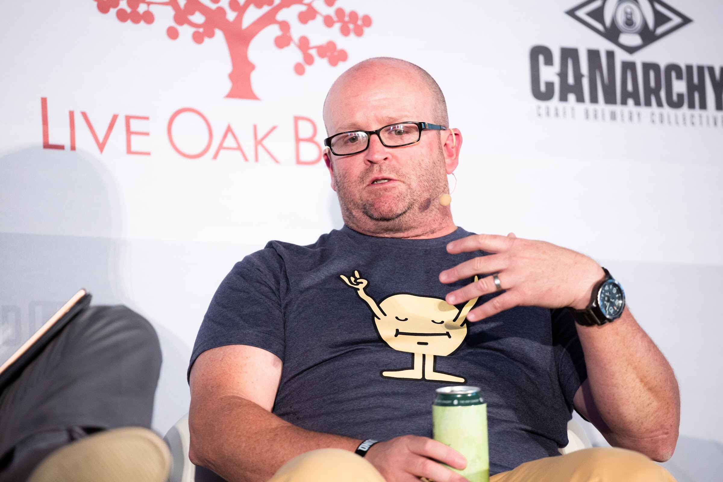 Bald man with glasses in a navy T-shirt seated on stage holding a can and gesturing.