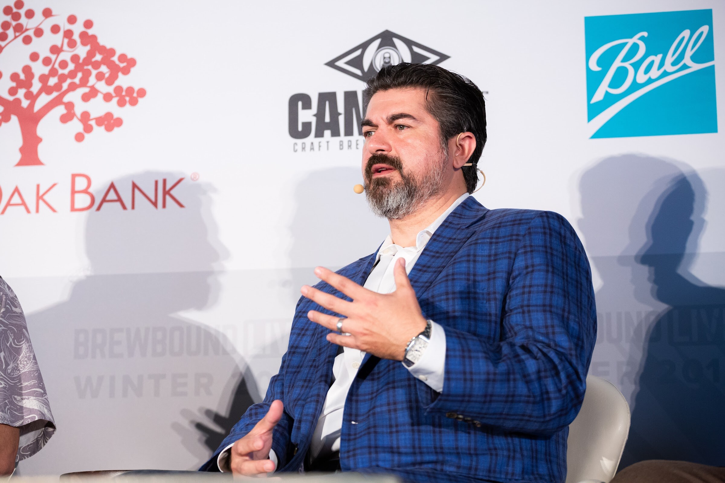 Man with a beard in a blue plaid blazer gesturing with both hands while seated in front of a sponsor backdrop.