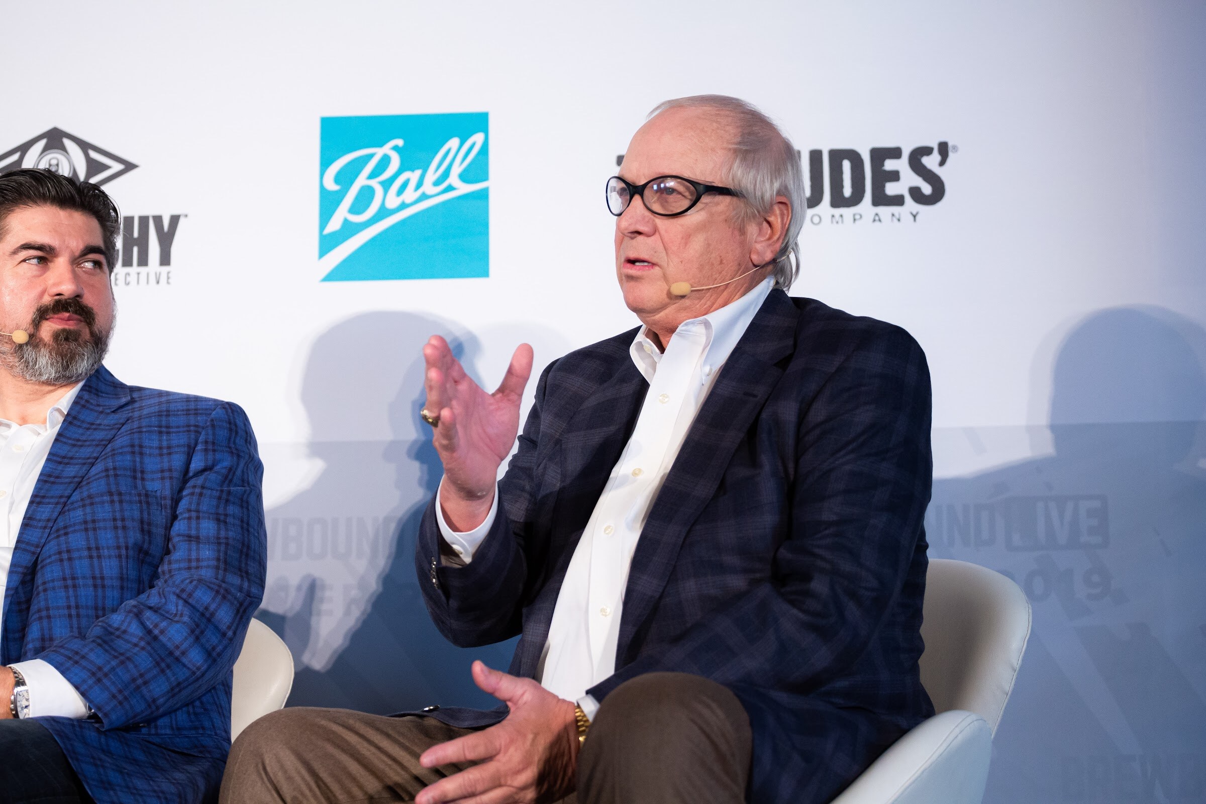 Two men in blazers seated on stage, one in glasses speaking with a raised hand.