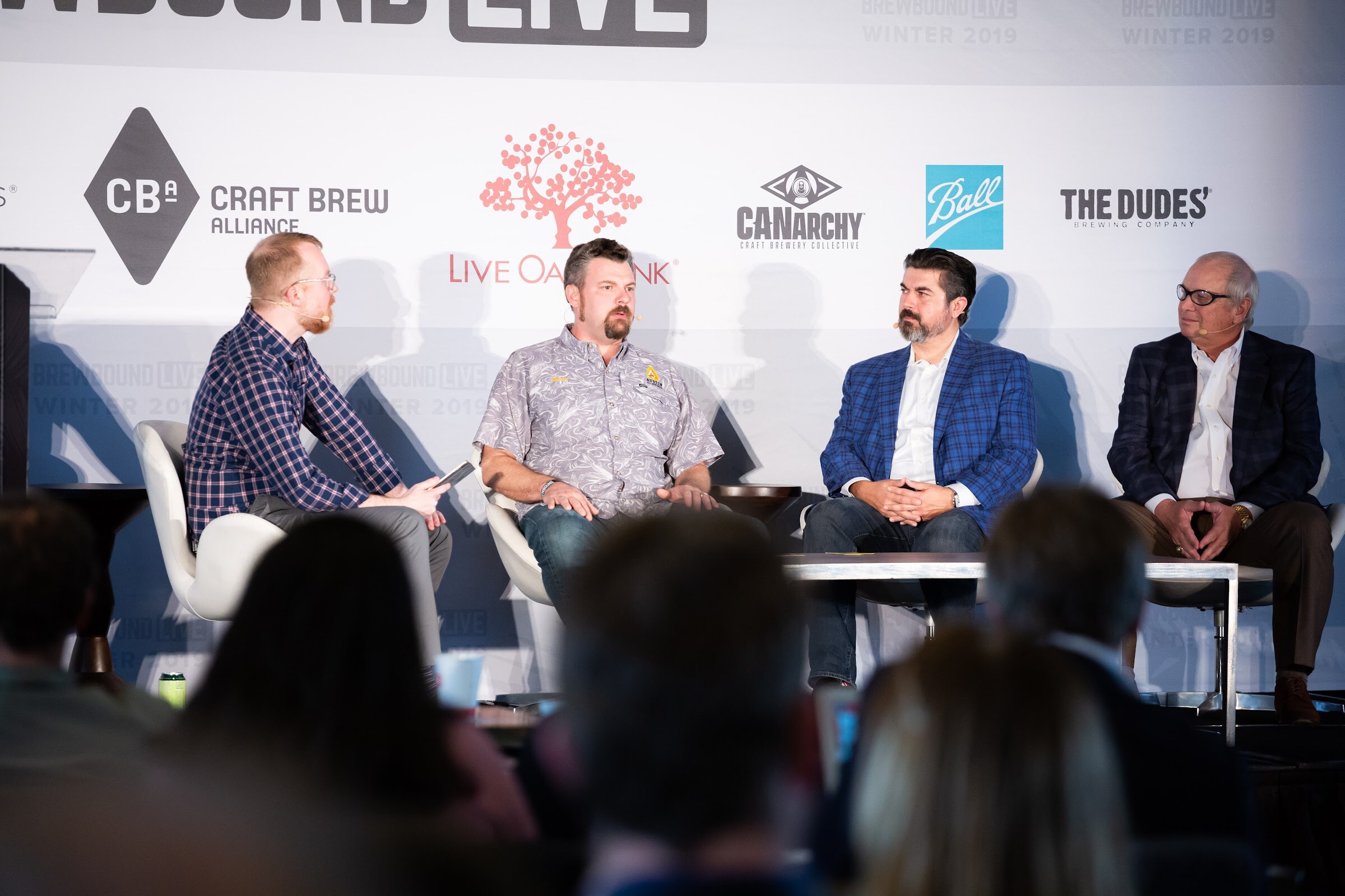 Four seated men on a stage in front of a sponsor wall with audience heads in the foreground.