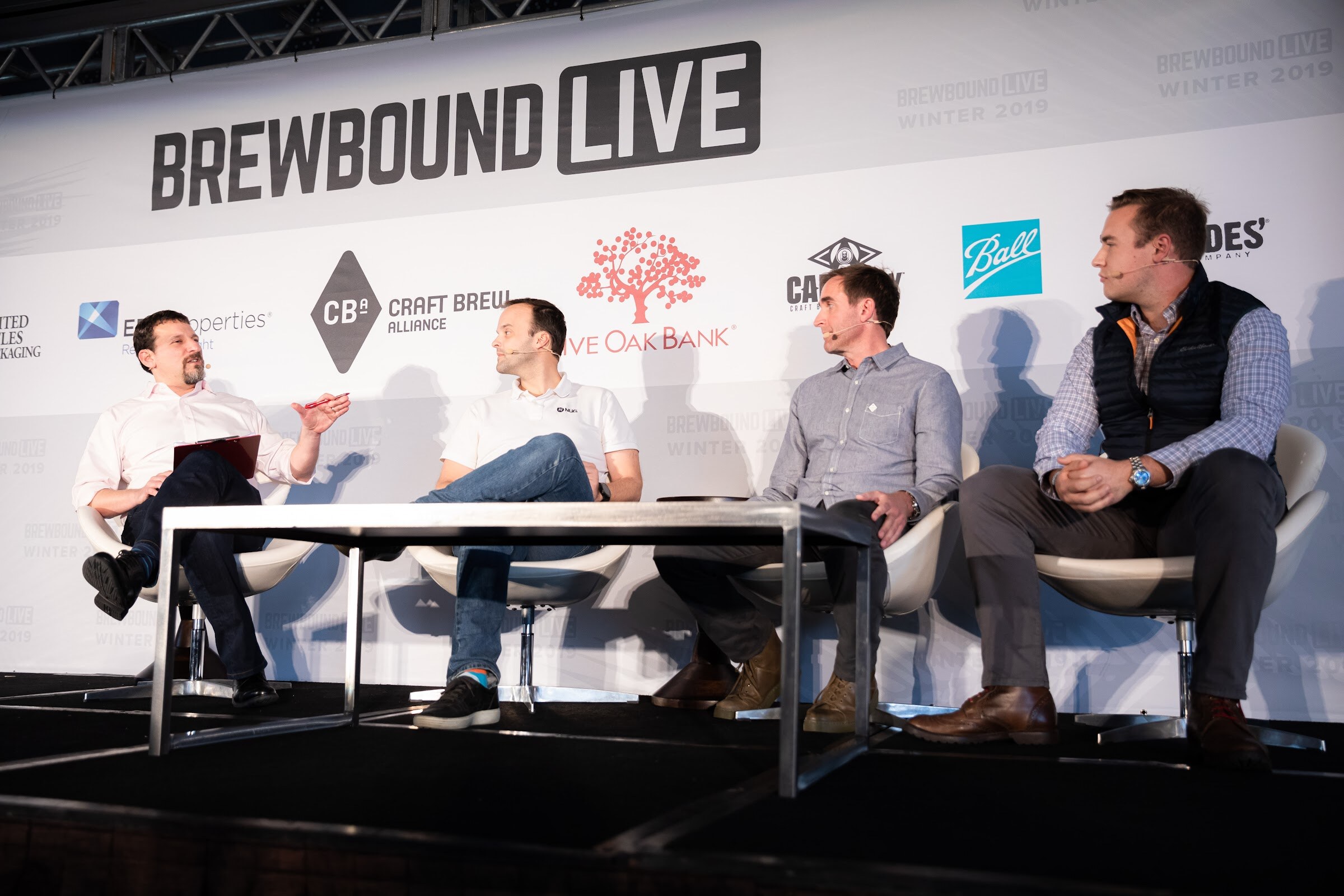 Four men seated in white chairs at a glass-top table on stage in front of the Brewbound Live backdrop.