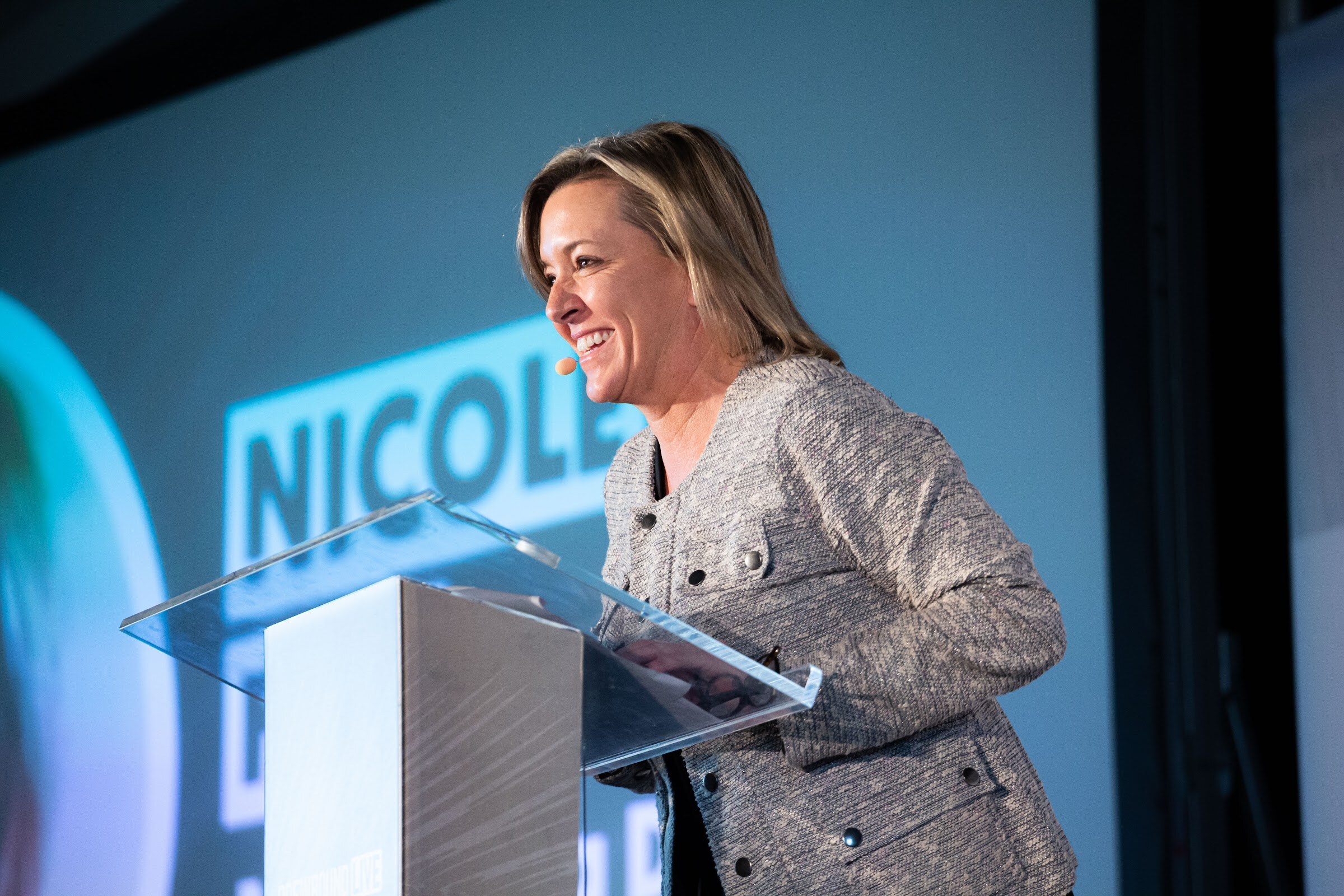 Woman in a gray tweed jacket at a glass lectern in front of a blue screen reading NICOLE.