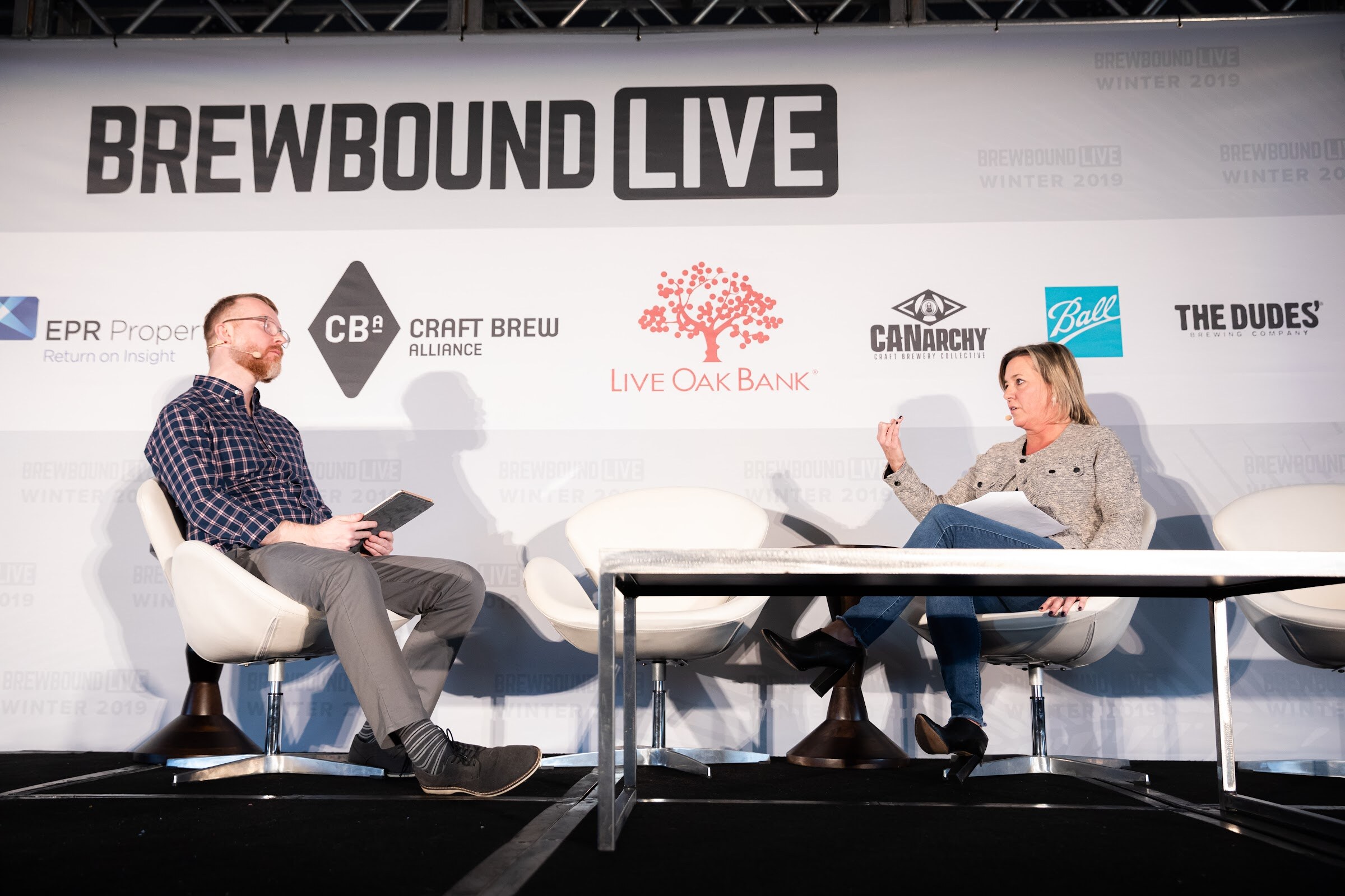Man in a plaid shirt and a woman in a gray jacket seated on stage in front of the Brewbound Live backdrop.