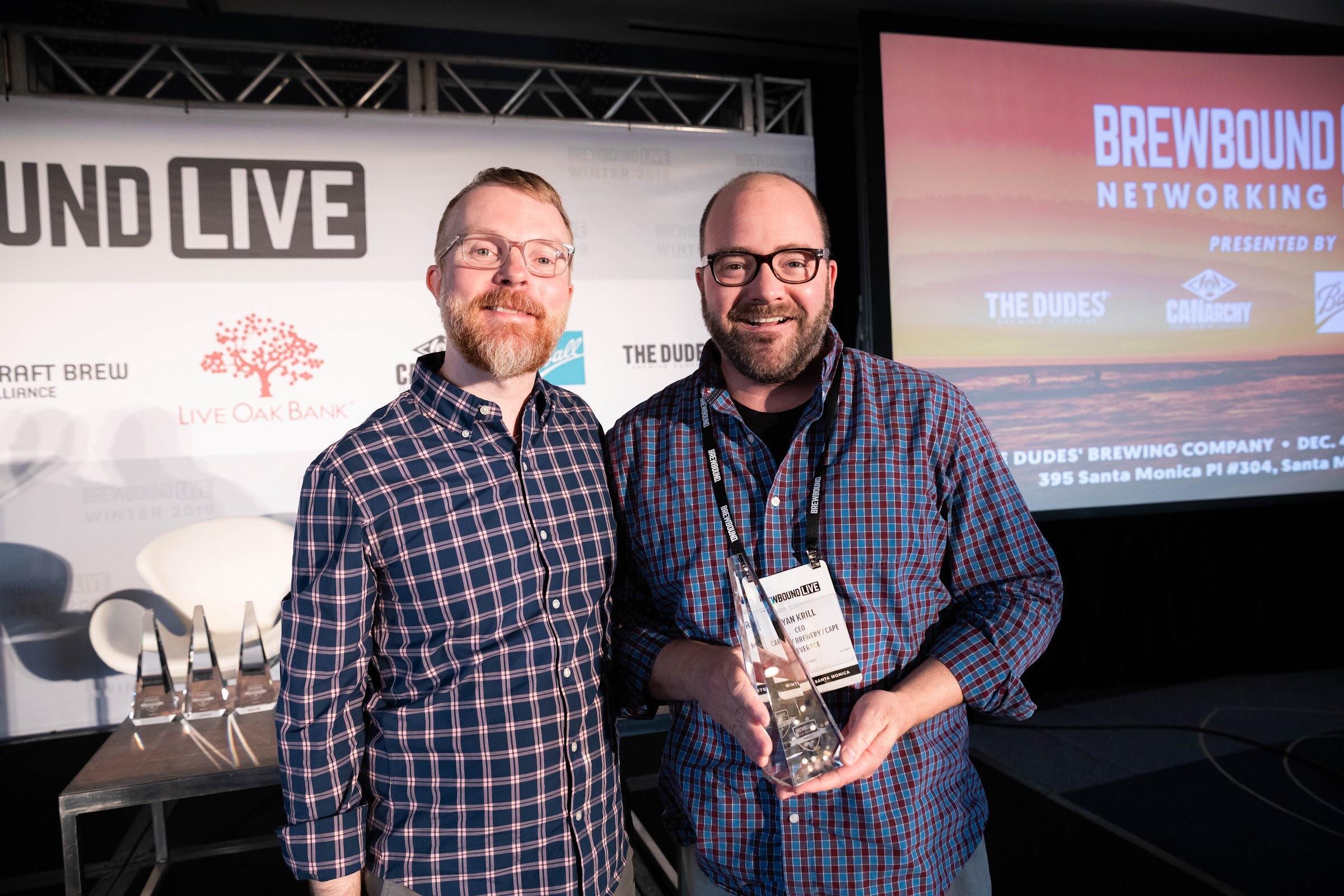Two men in plaid shirts, one holding a glass trophy, in front of the Brewbound Live backdrop and a screen advertising the networking party.