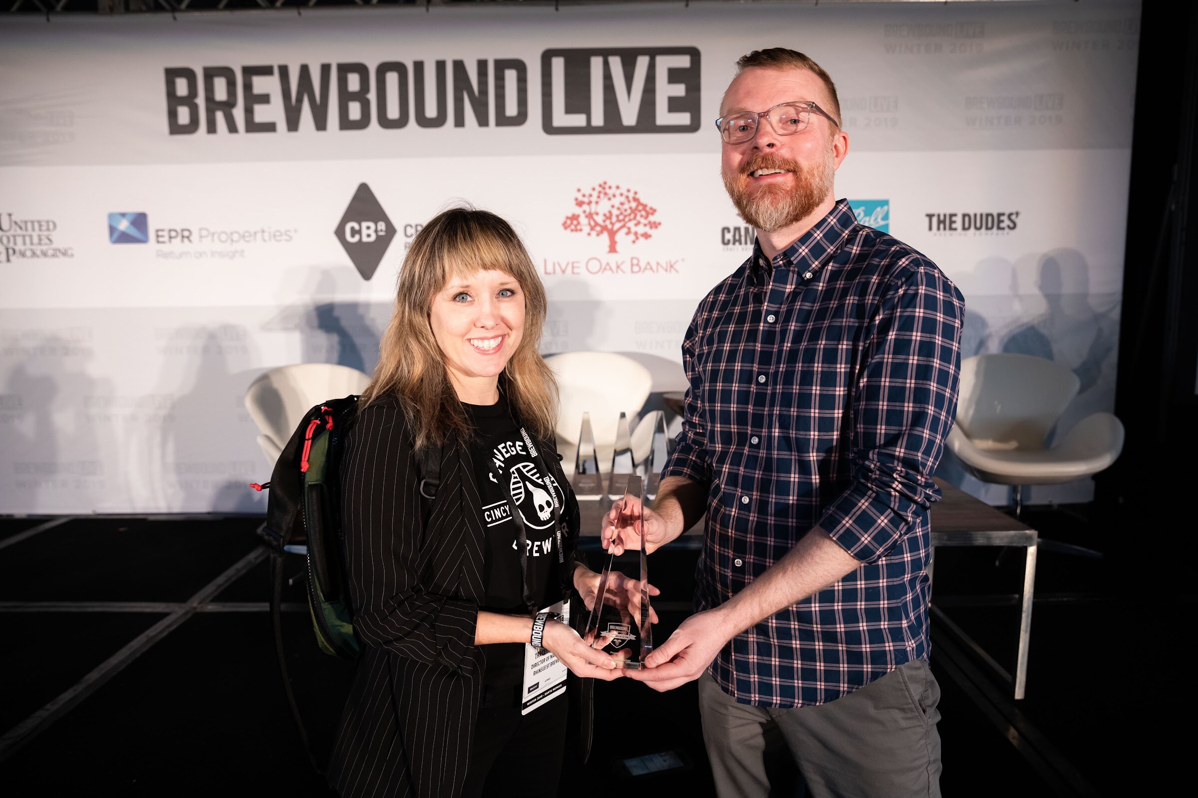 Woman in a black pinstripe jacket and a man in a plaid shirt holding a glass award on stage.