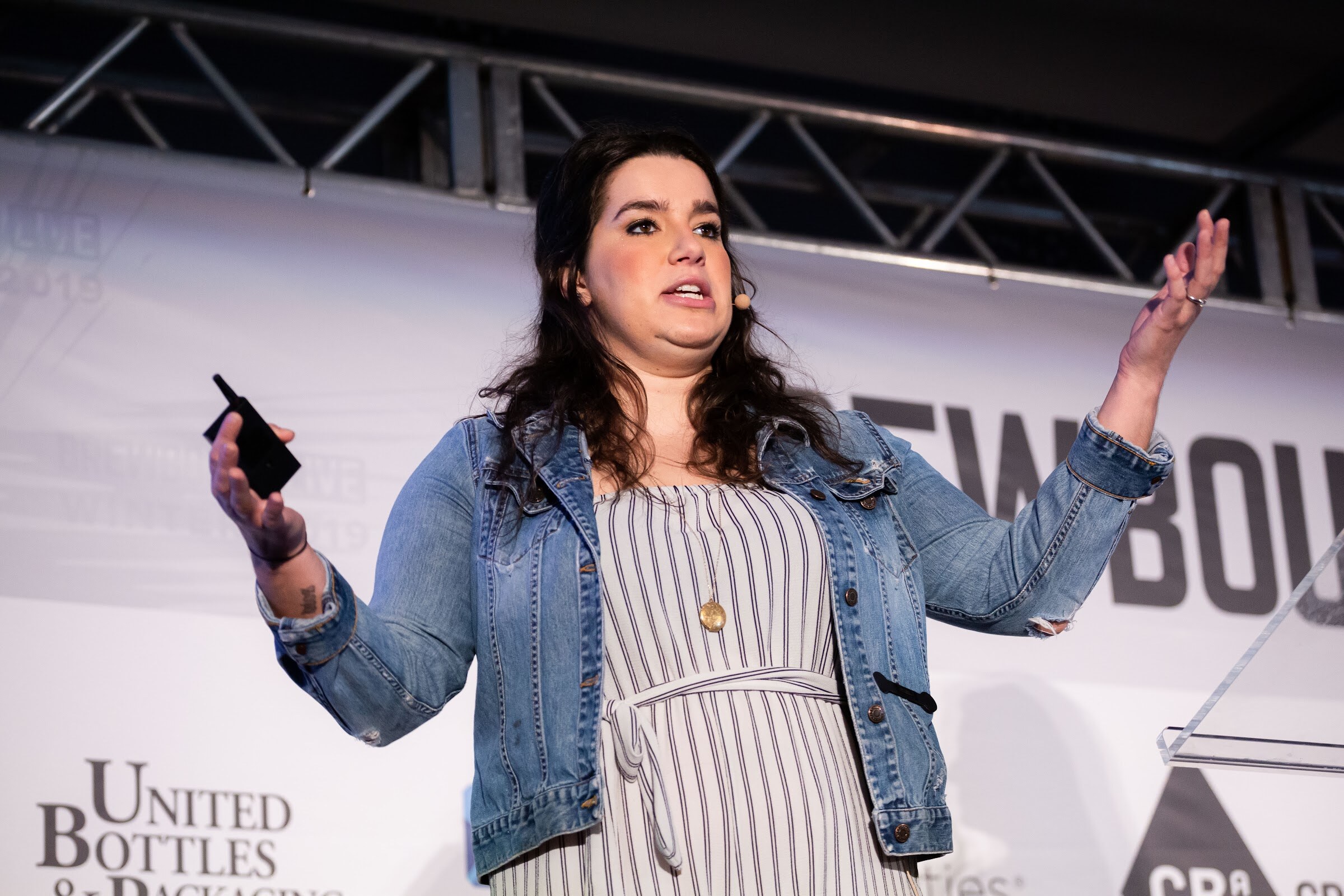 Woman in a denim jacket and striped dress on stage with both arms raised, holding a clicker.