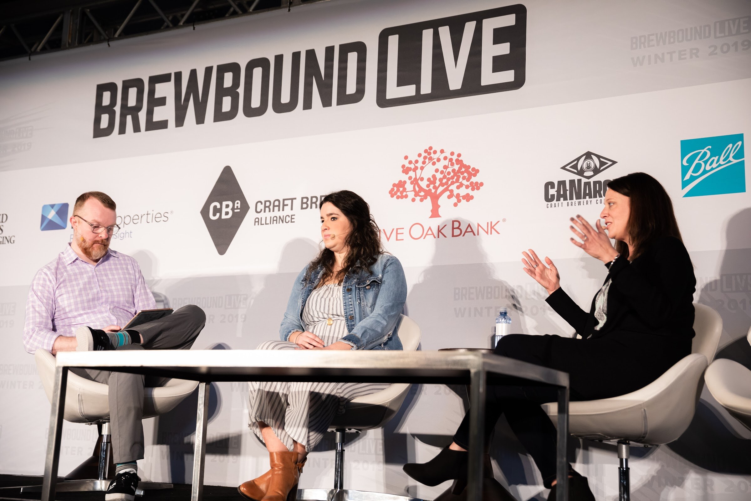 Three people seated at a metal table on stage: a man in a lavender shirt, a woman in a denim jacket and a woman in black.