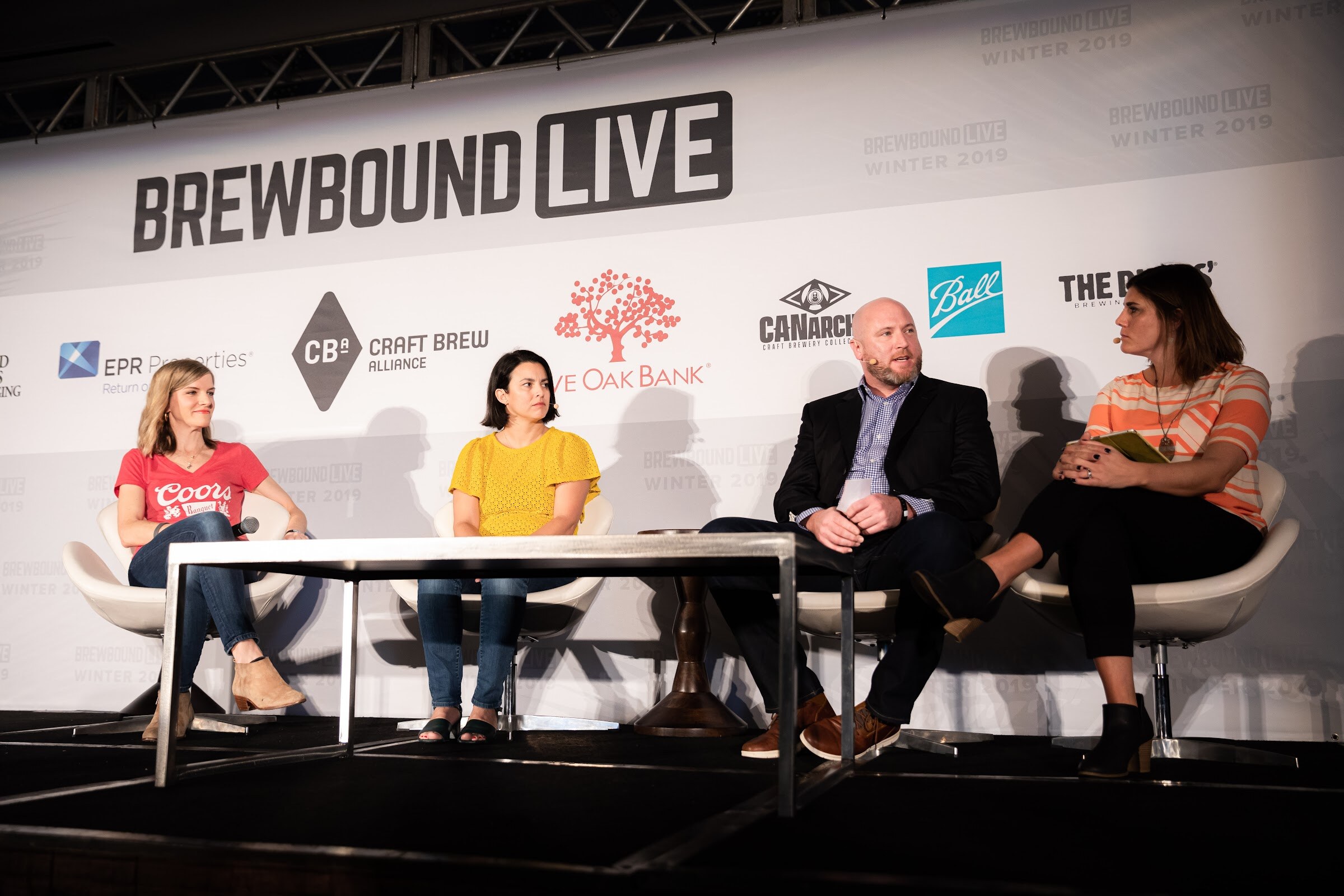 Four seated people on stage at a long table; one woman wears a red Coors Banquet T-shirt and another a yellow top.