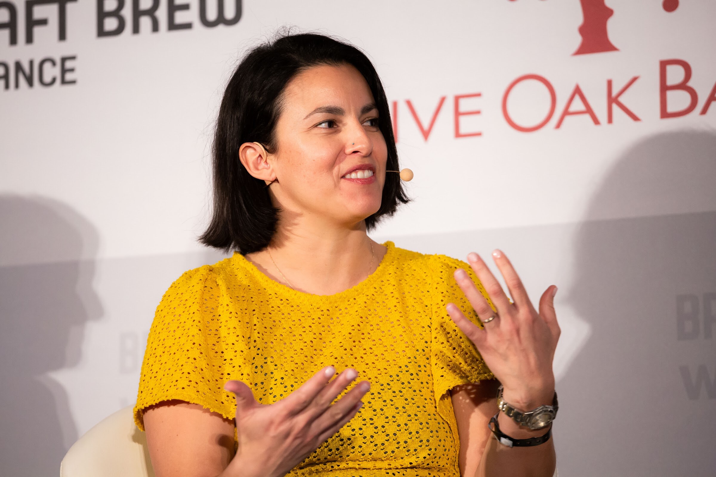 Woman in a yellow short-sleeve top gesturing with both hands, seated in front of a Live Oak Bank banner.
