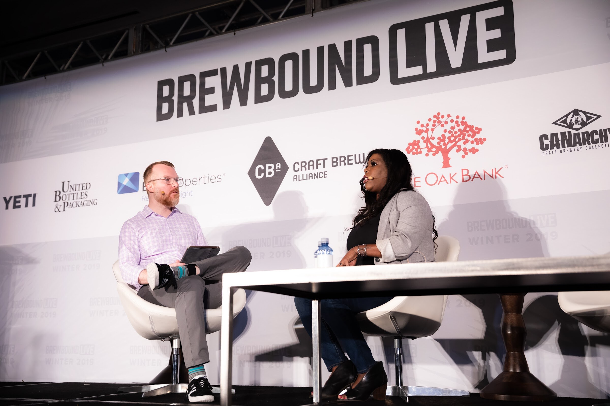 Man in a lavender shirt and a woman in a gray blazer seated on stage in front of the sponsor backdrop.