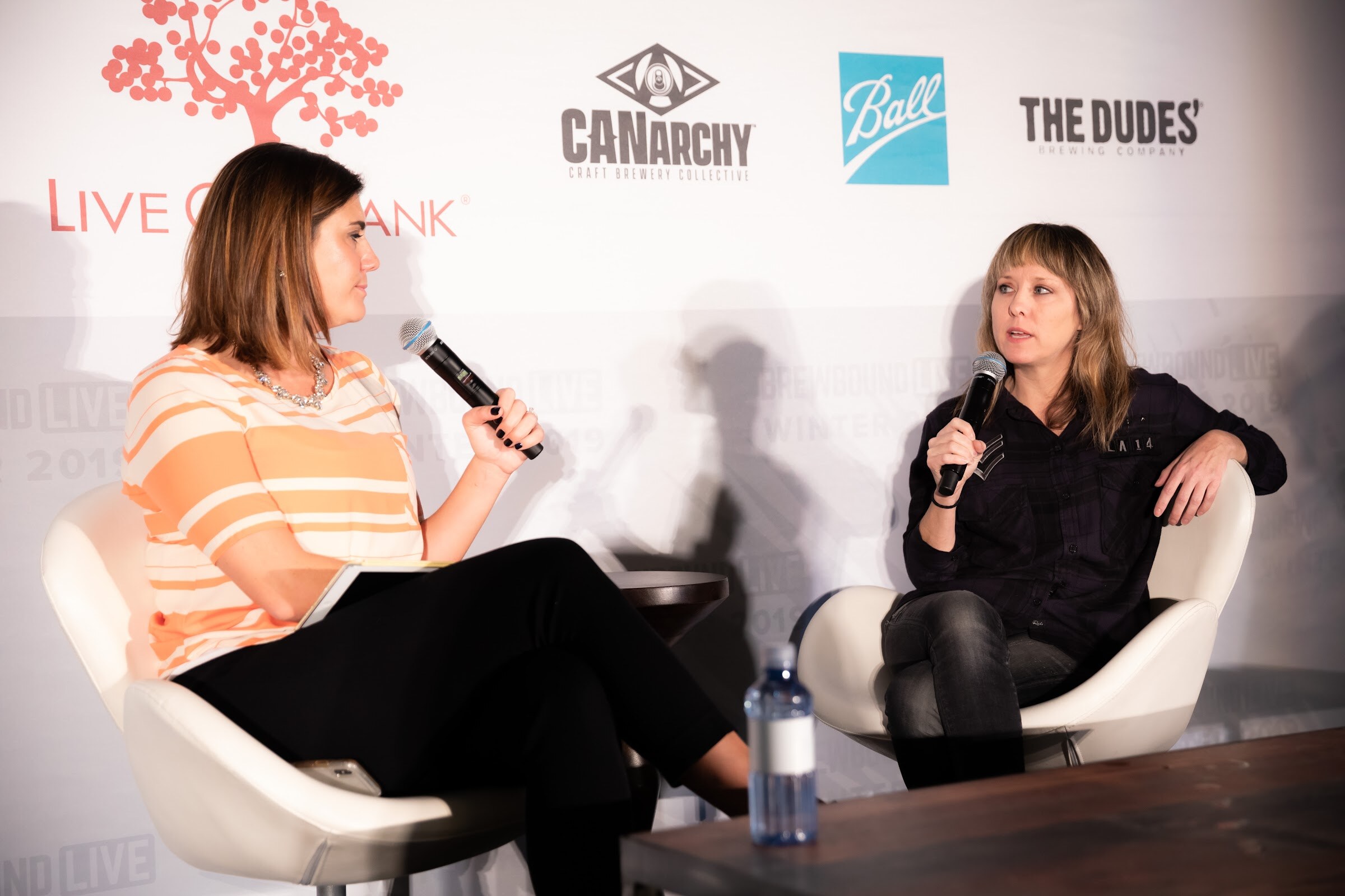 Woman in an orange striped top holding a microphone facing a woman in black seated in a white chair.