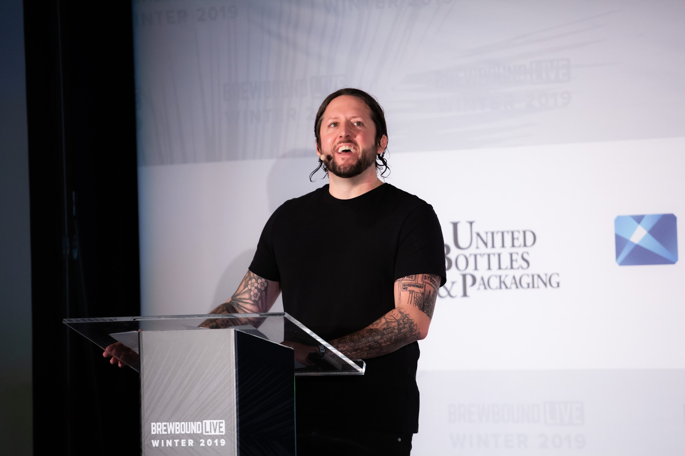 Tattooed man in a black T-shirt smiling at a glass lectern with a Brewbound Live Winter 2019 sign.