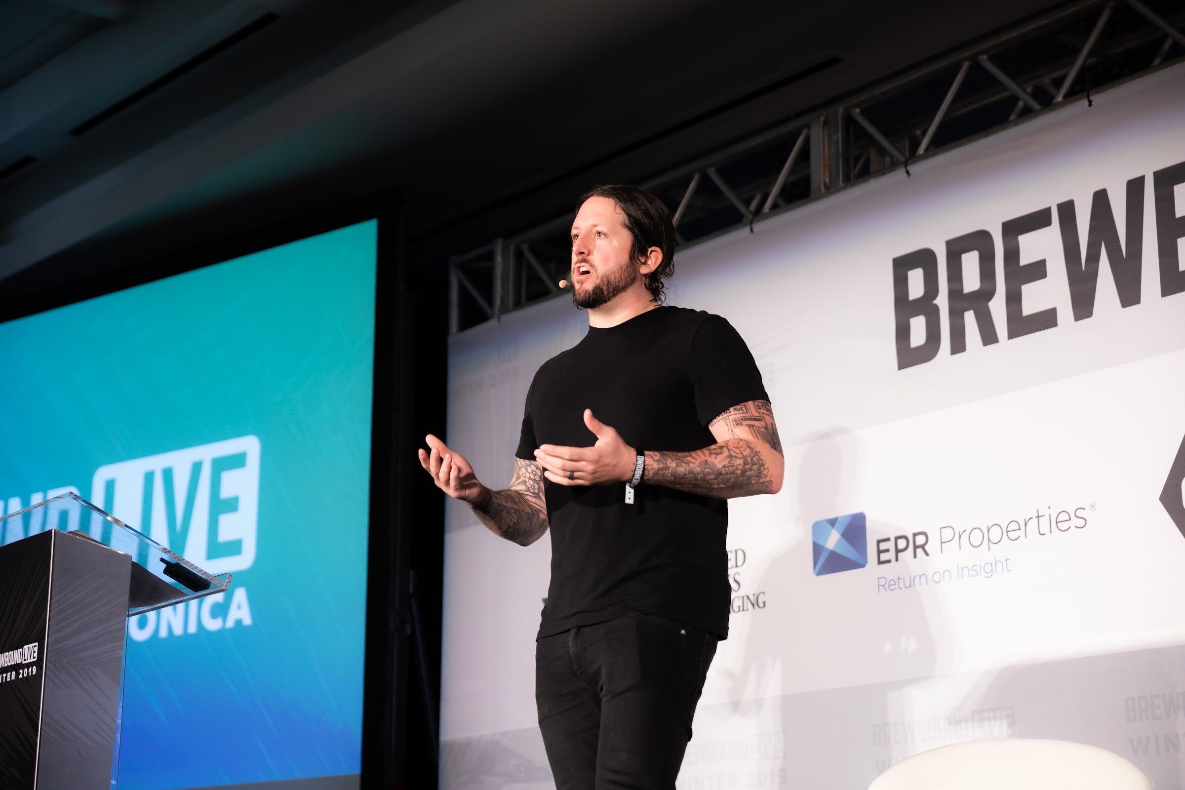 Man in a black T-shirt with tattooed arms speaking on stage beside a blue screen.