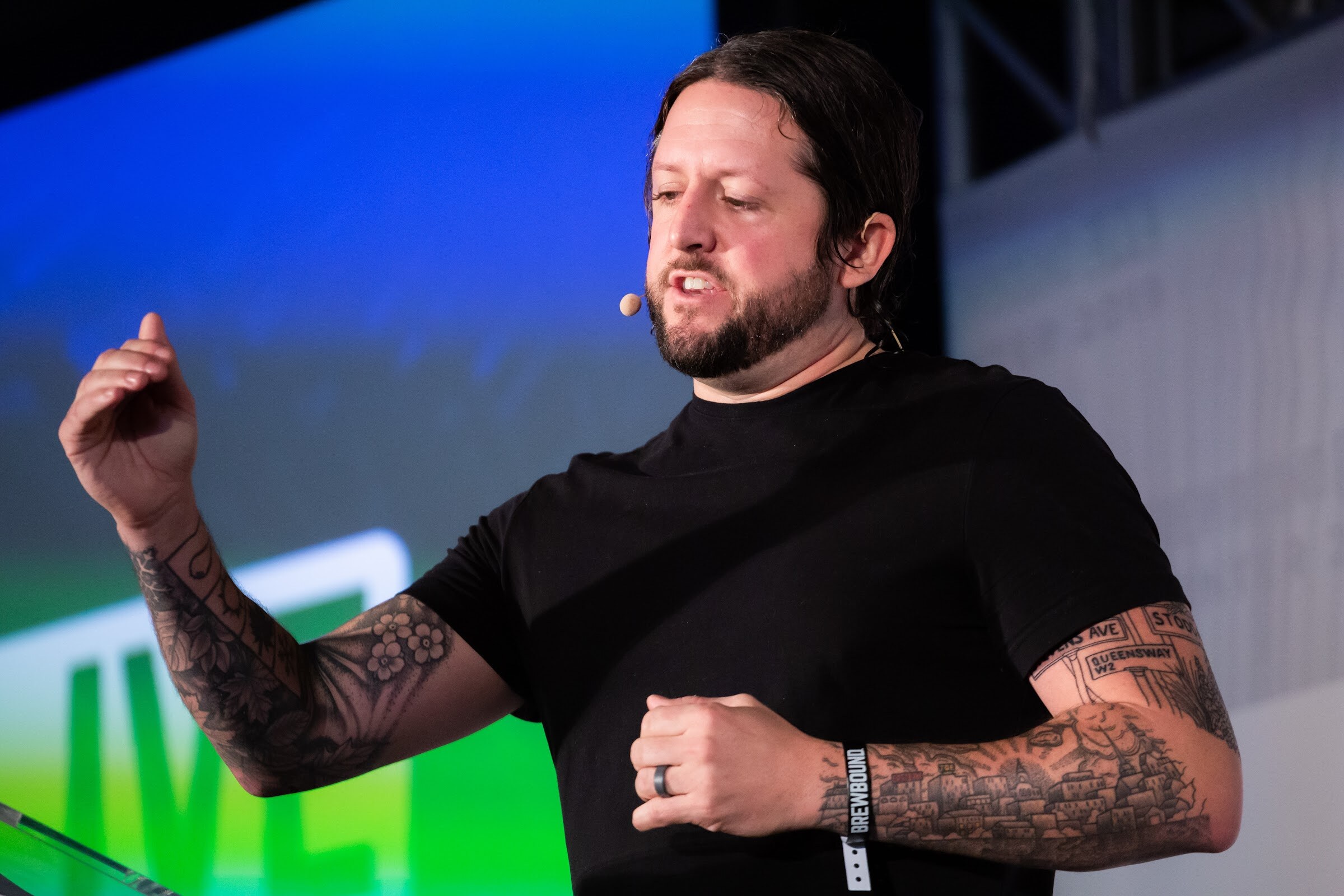 Tattooed man in a black T-shirt speaking with one hand raised in front of a blue and green screen.