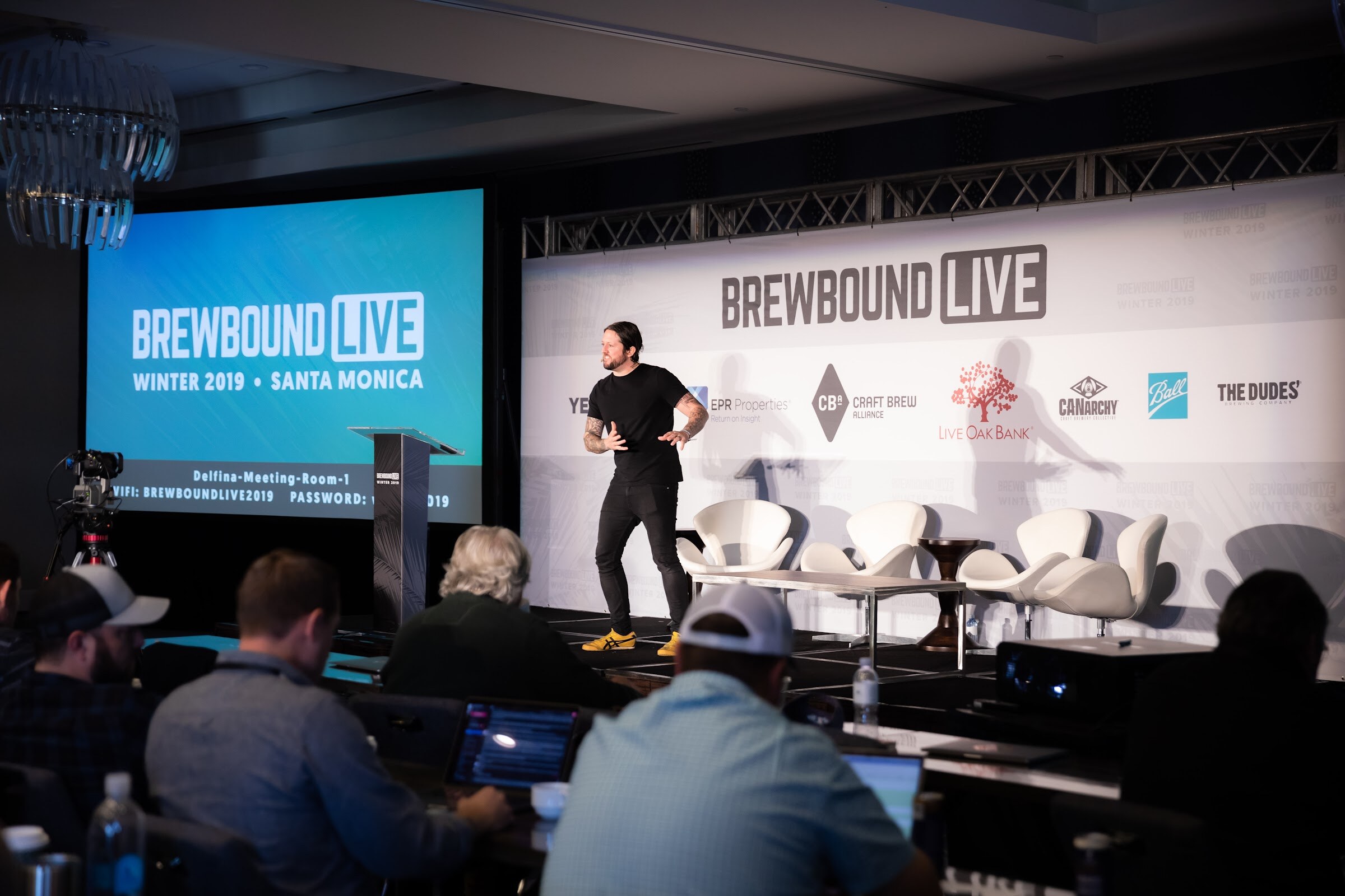 Wide view of a stage with a man in black, a screen reading Brewbound Live Winter 2019 Santa Monica, and attendees with laptops in the foreground.