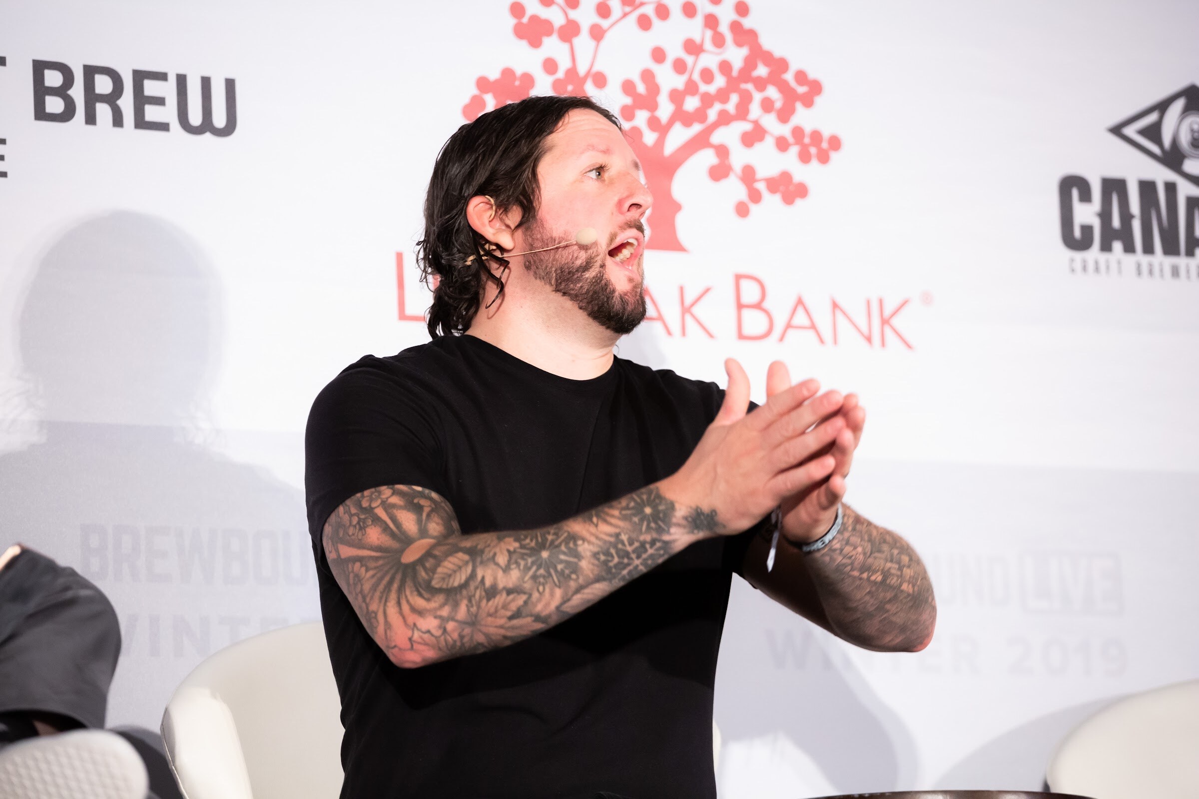Man in a black T-shirt with tattooed forearms clasping his hands, seated in front of a Live Oak Bank banner.