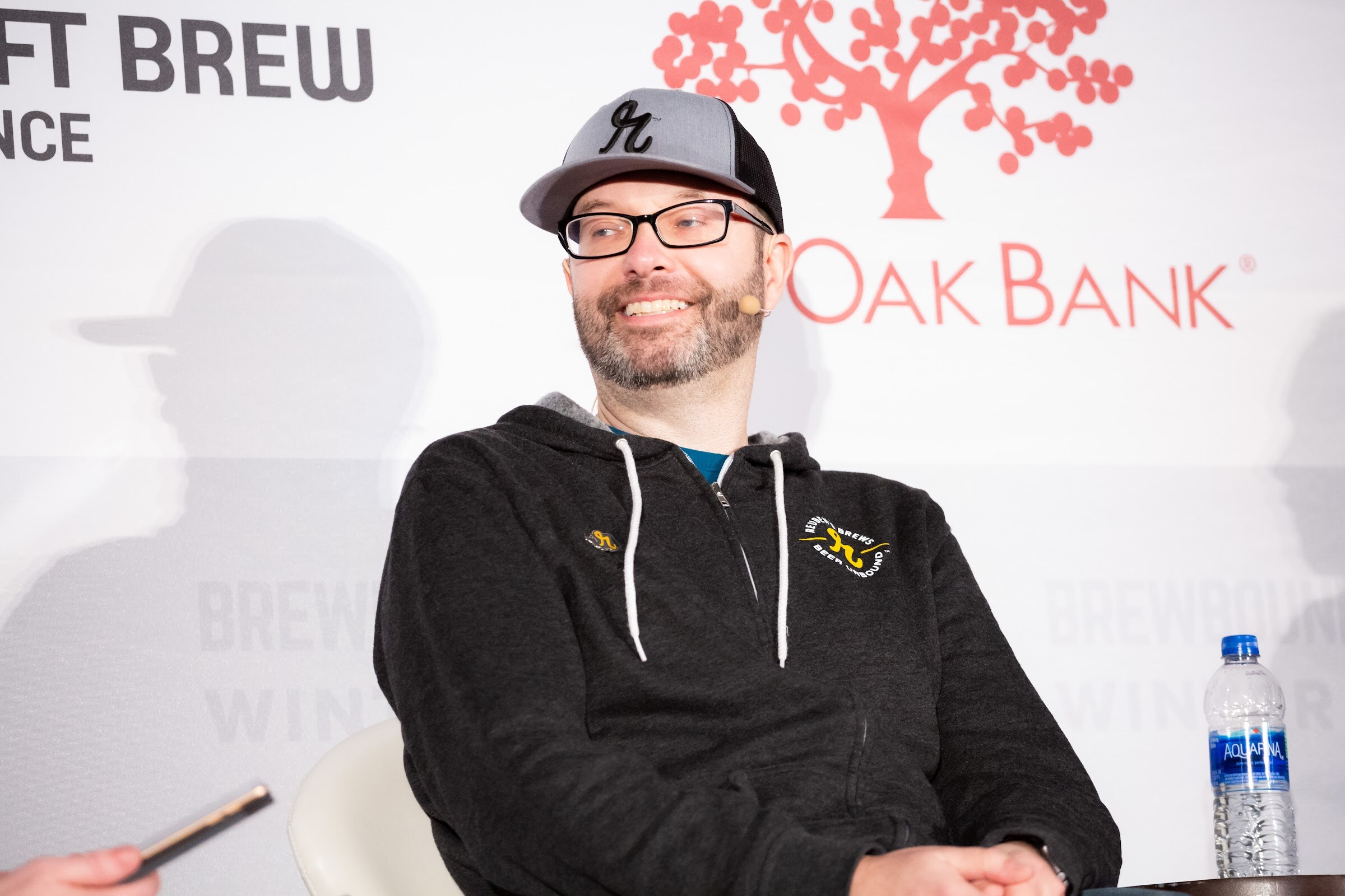 Man with glasses and a gray cap in a black zip hoodie smiling, seated on stage.
