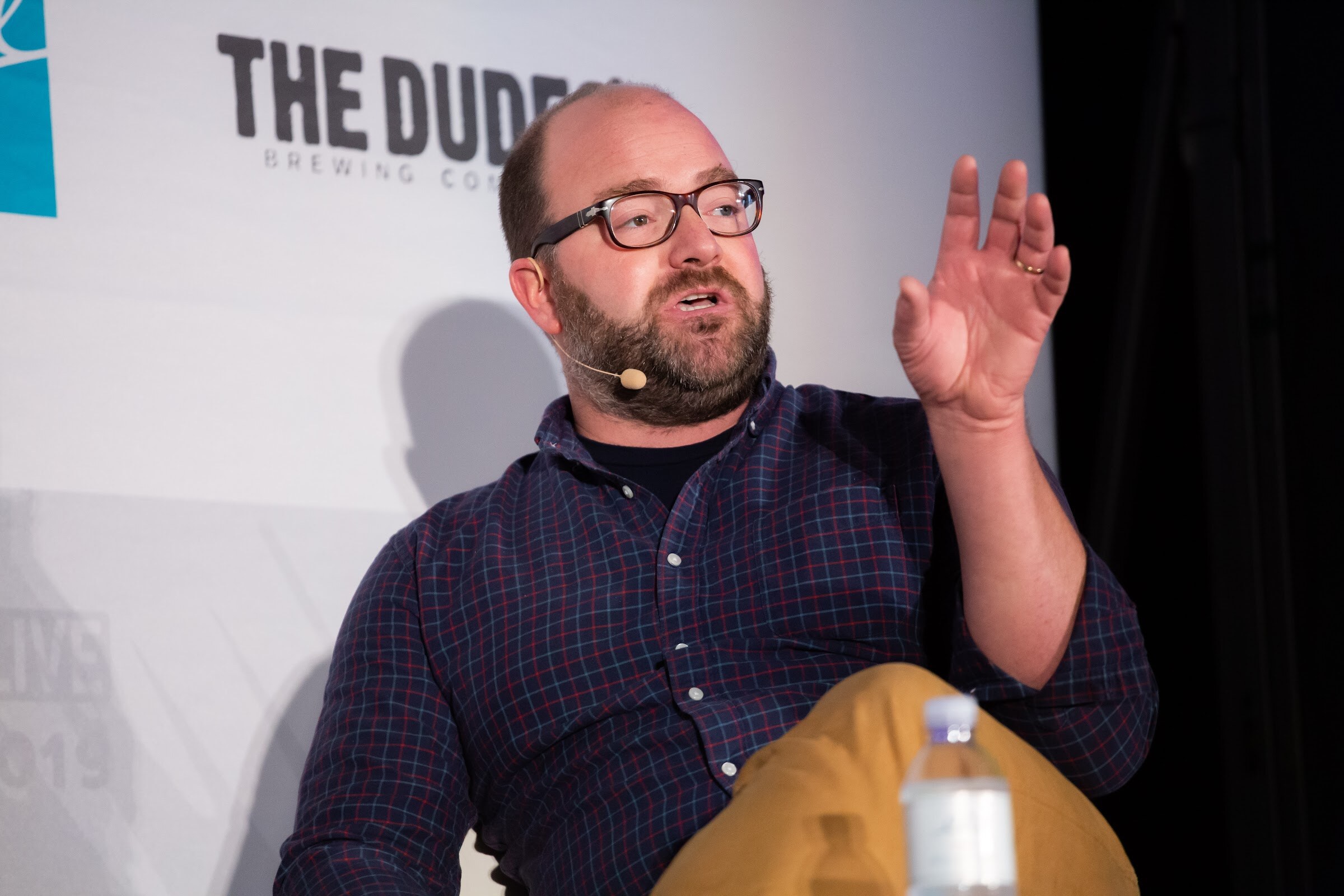 Bearded man in glasses and a navy plaid shirt raising one hand while seated on stage.