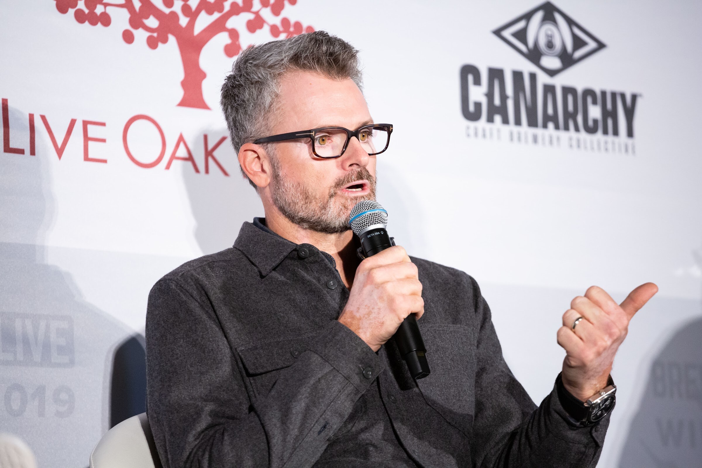Man with glasses and graying hair in a charcoal shirt holding a microphone and pointing his thumb over his shoulder.
