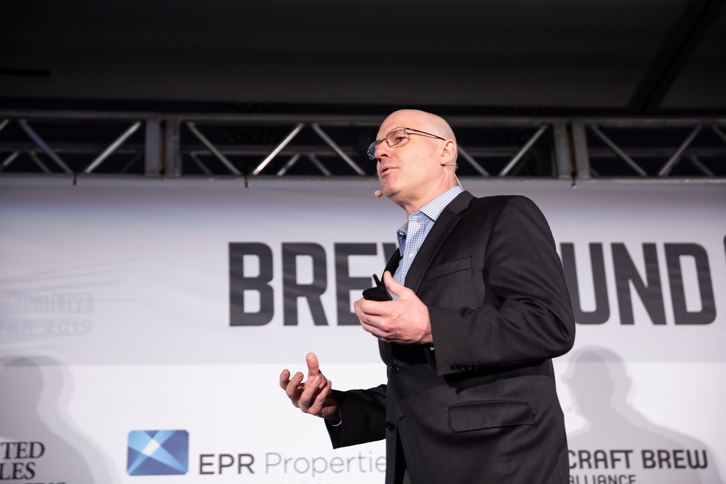 Bald man with glasses in a dark blazer on stage gesturing with one hand and holding a clicker.