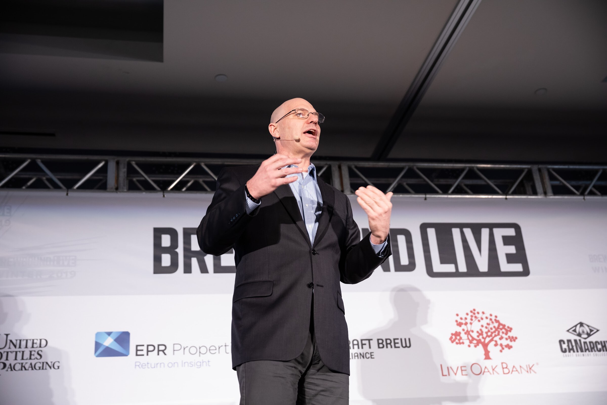 Bald man in glasses and a dark blazer with both hands raised in front of the Brewbound Live backdrop.