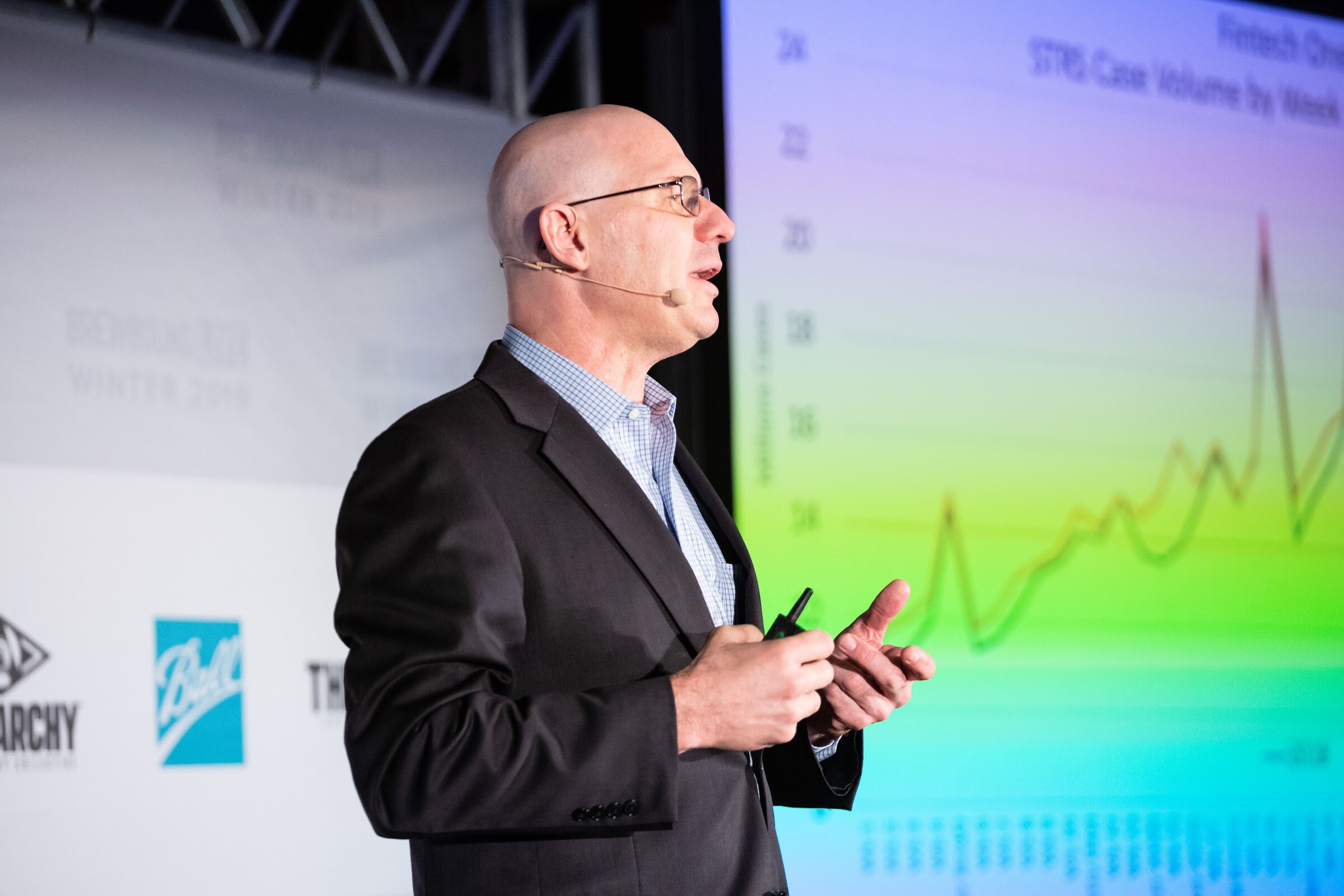 Man in a dark blazer standing beside a projected line chart of case volume by week.