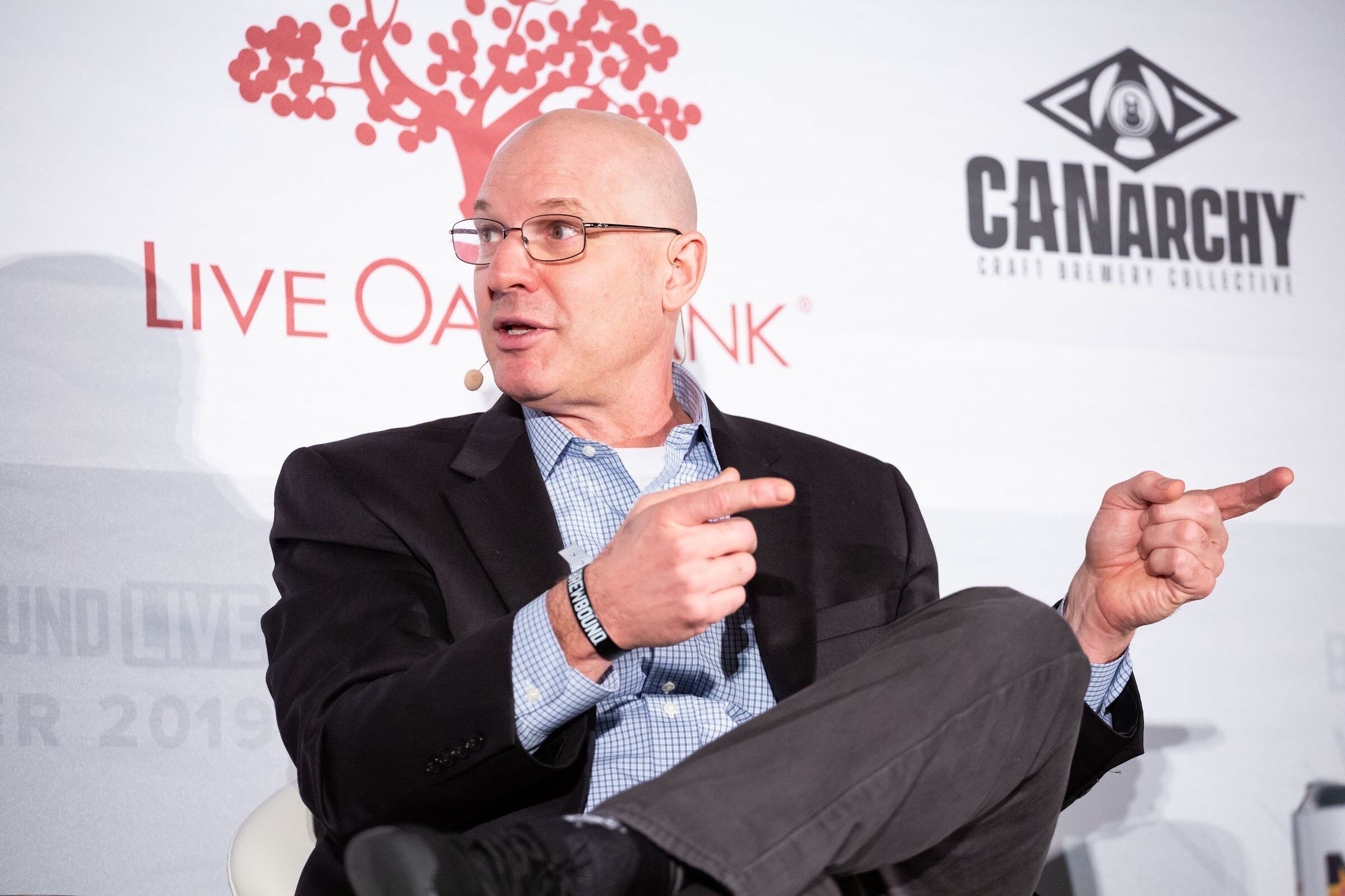 Bald man in glasses and a dark blazer seated with both index fingers pointed.