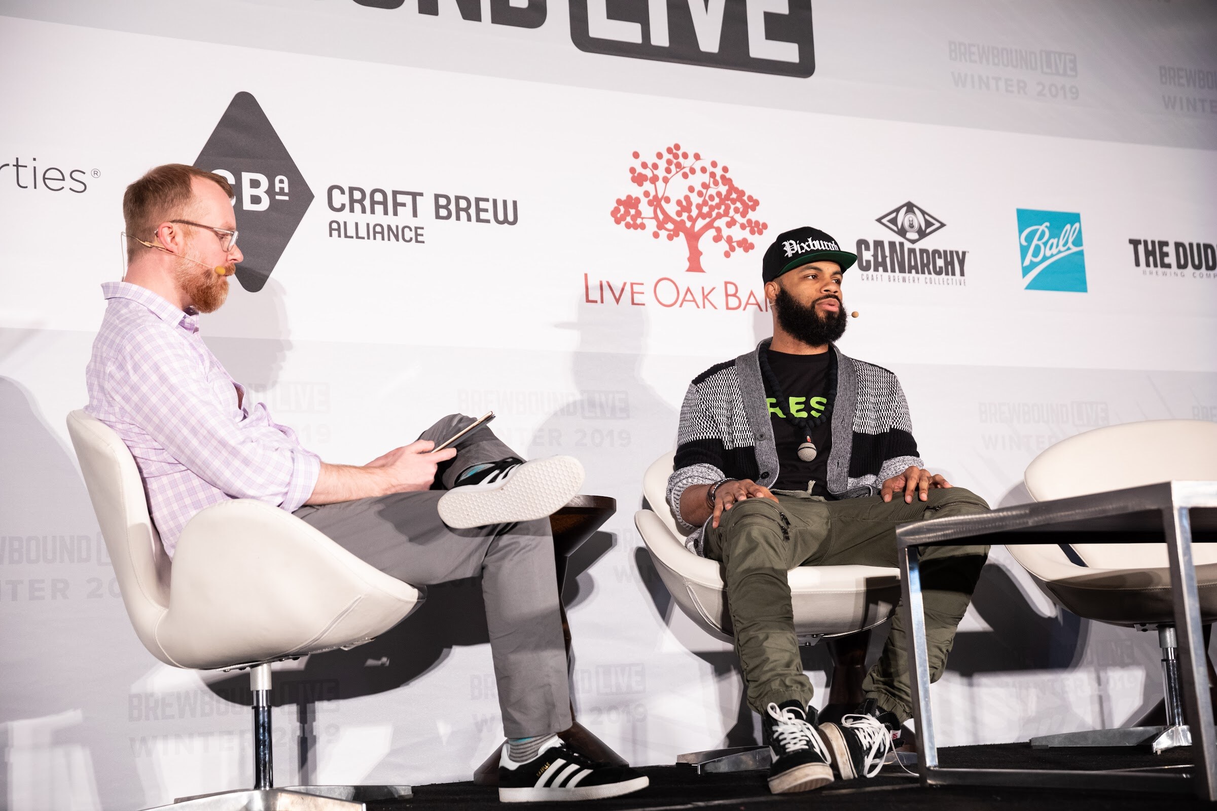 Man in a lavender checked shirt seated beside a bearded man in a black Pixburgh cap and gray cardigan.