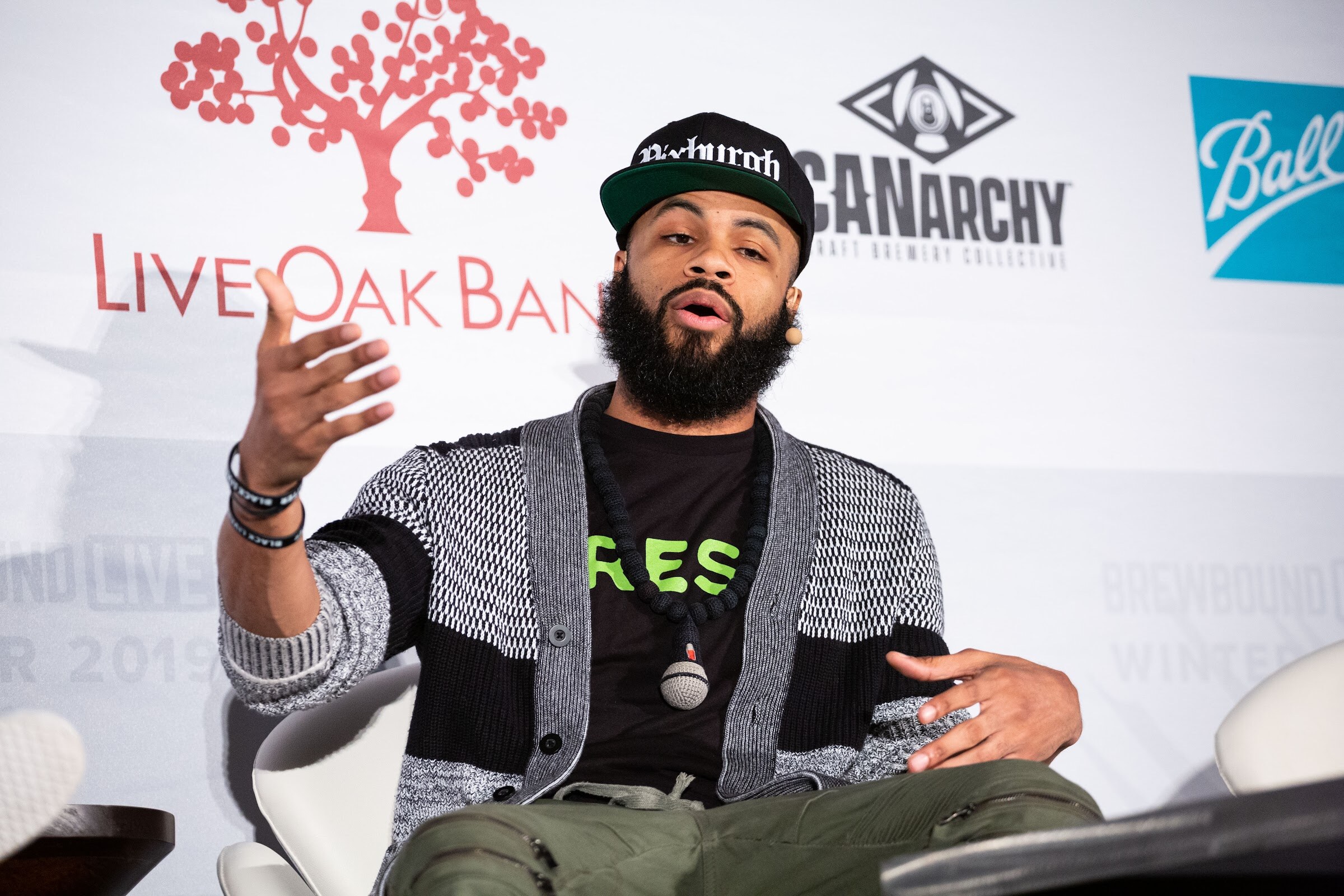 Bearded man in a black cap and gray cardigan raising one hand while speaking, with a Live Oak Bank banner behind him.