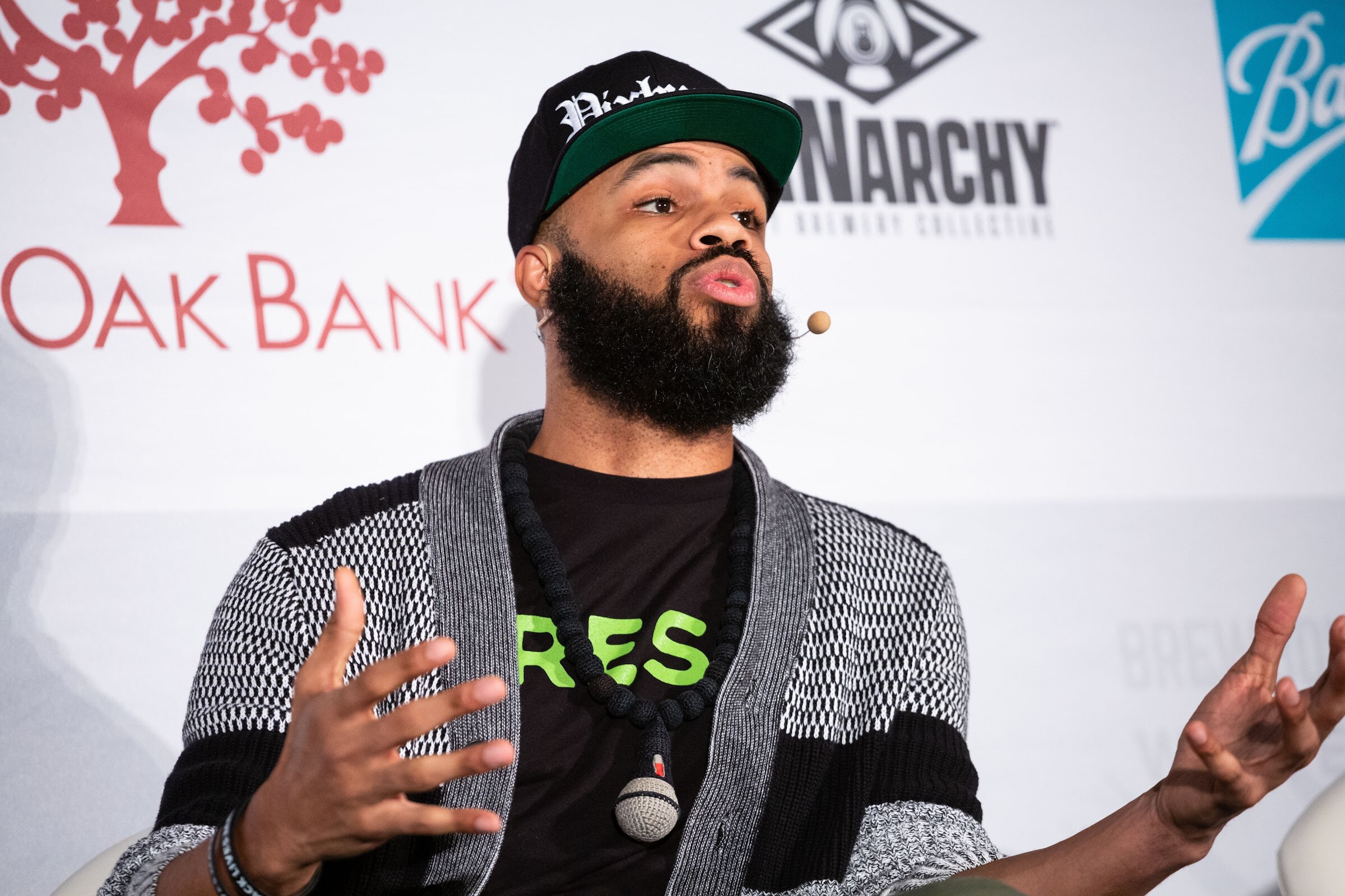 Bearded man in a cap and gray cardigan with both hands open, in front of a Live Oak Bank banner.
