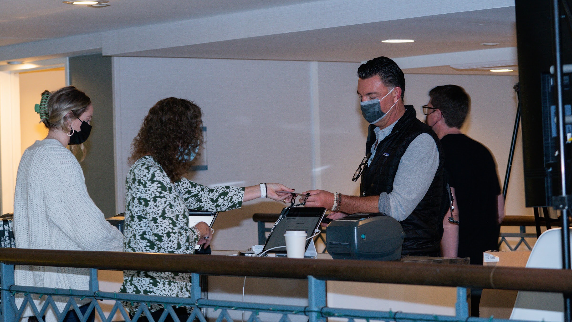 Two women and a man in a vest at a registration counter with a laptop and badge printer, the man receiving a lanyard.