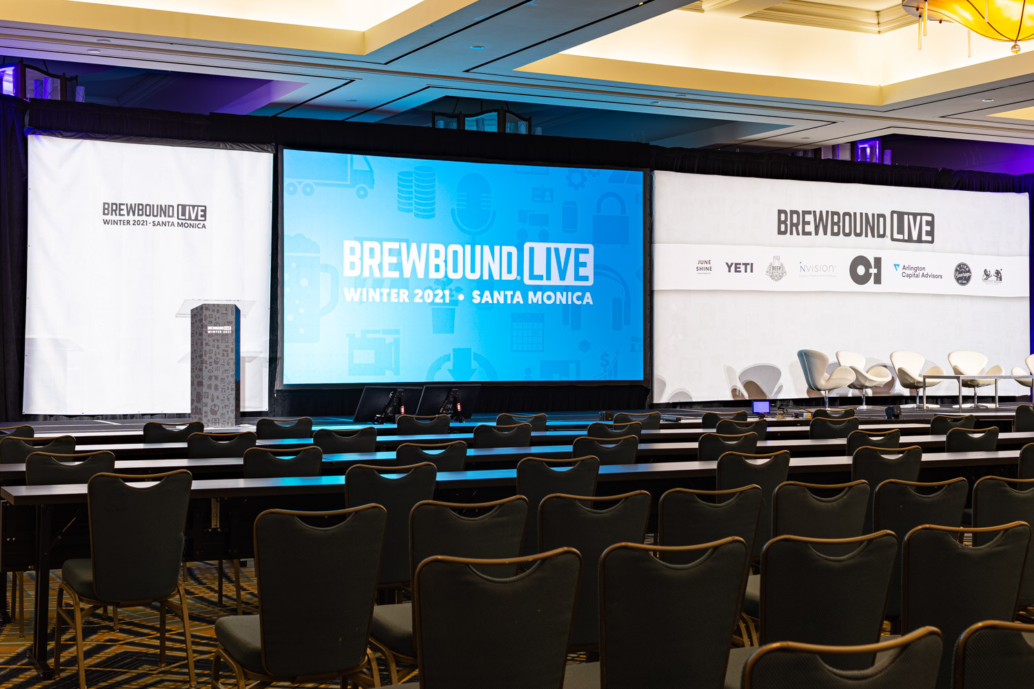 A ballroom with rows of tables and chairs facing a stage with a blue Brewbound Live Winter 2021 Santa Monica screen, a podium and white chairs.