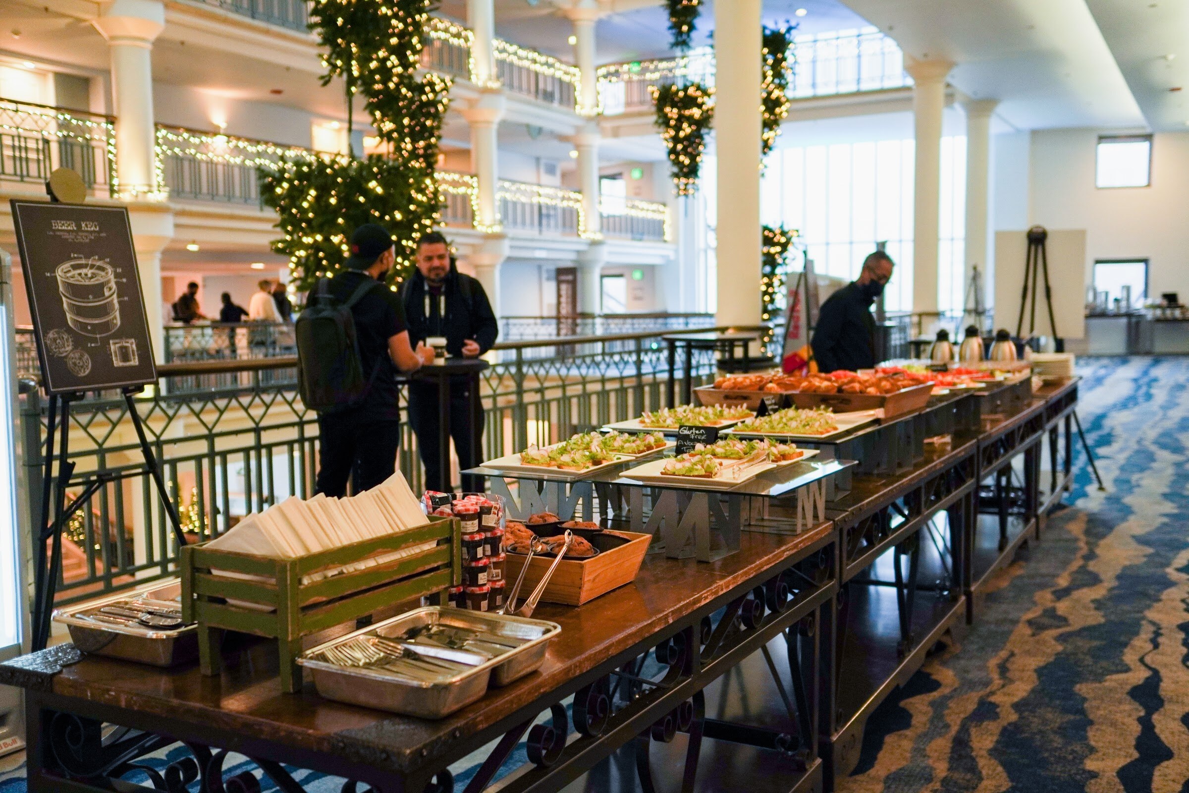 A long buffet table of breakfast dishes stands beneath a holiday-lit atrium as a few attendees stand nearby.