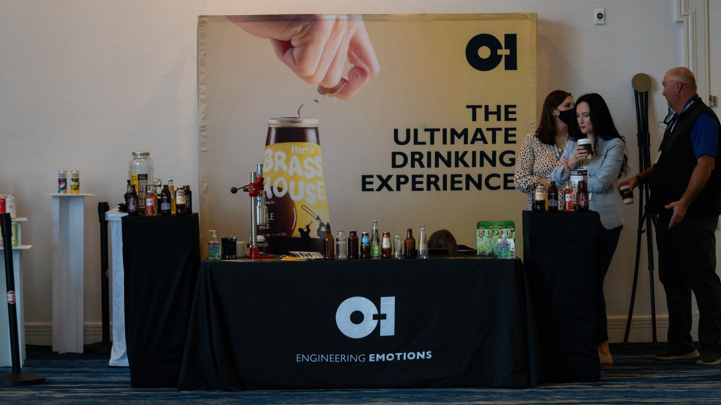 A black-draped table with glass bottles and a yellow O-I banner reading The Ultimate Drinking Experience, with three people standing beside it.