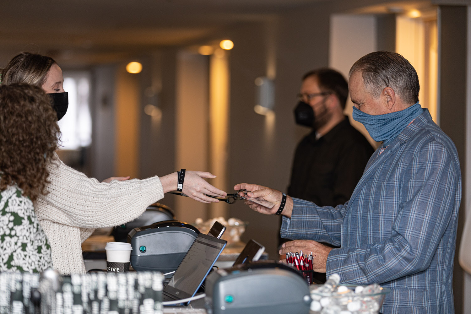 A woman in a cream sweater hands a lanyard to a man in a blue plaid shirt at a registration table with badge printers.