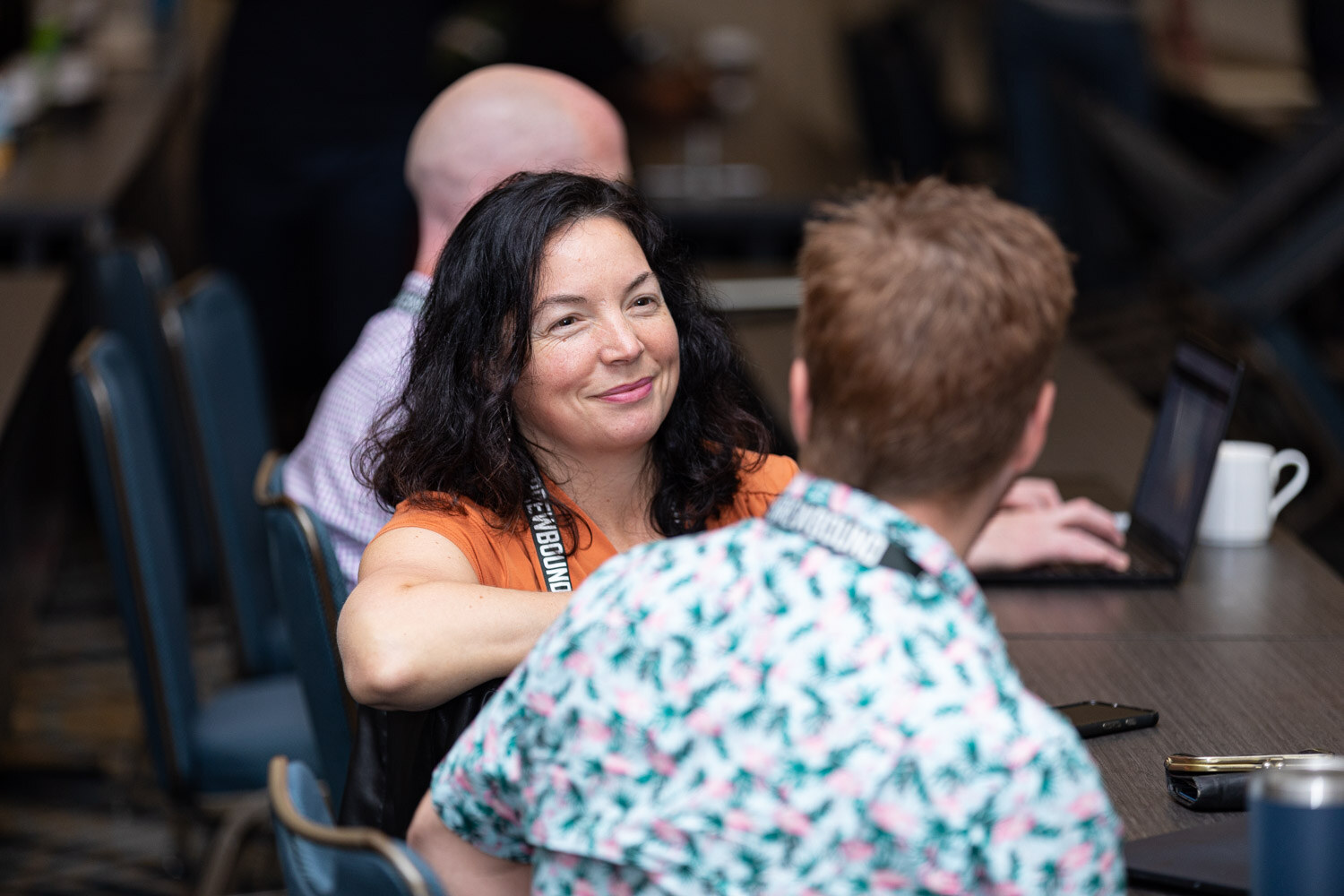 A smiling woman with dark hair in an orange top talks with a man in a teal patterned shirt at a table with a laptop and mug.