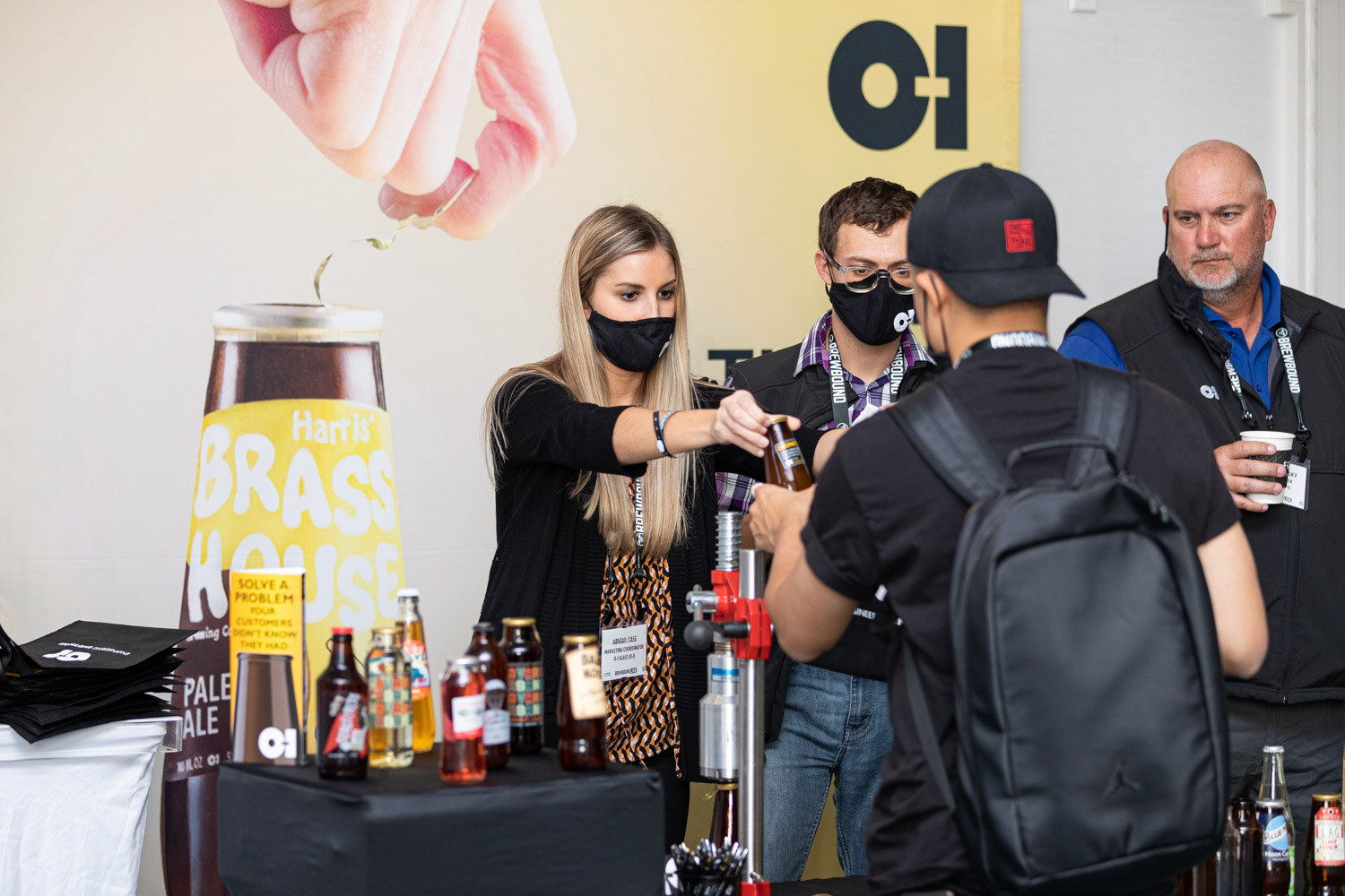 Four people stand at a black table of bottles beneath a large banner with the O-I logo and a Brass House bottle.