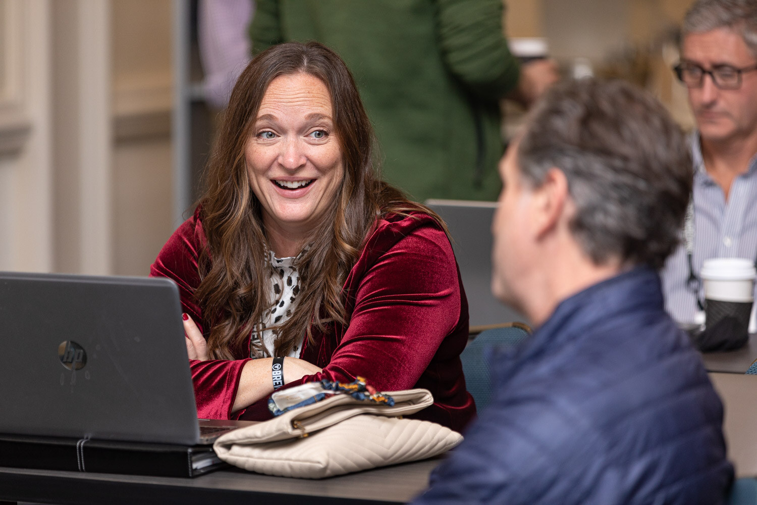 A woman in a red velvet jacket laughs at a table with a laptop while a man in a navy jacket faces her.