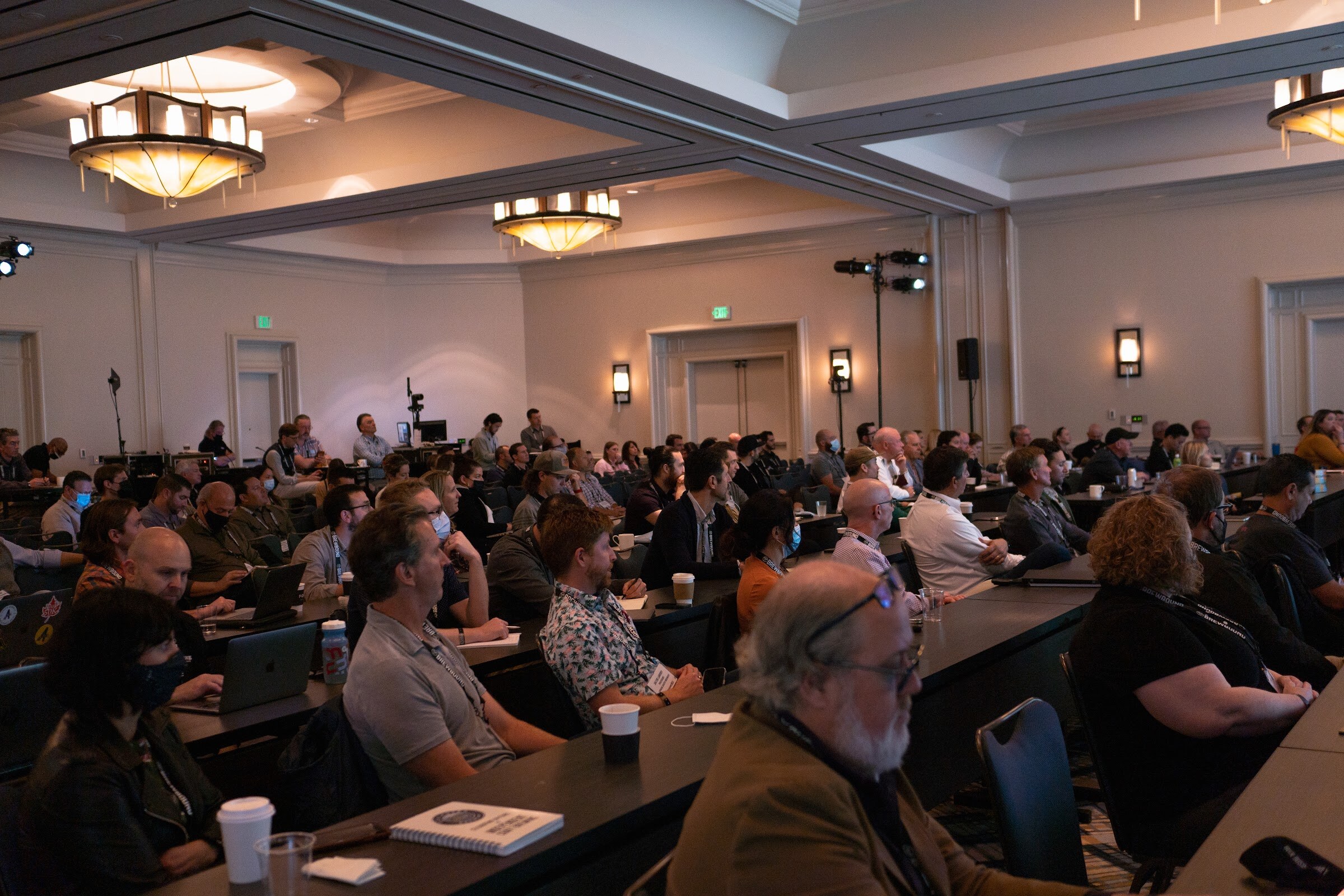 Rows of seated attendees with laptops and coffee cups face the stage in a hotel ballroom.