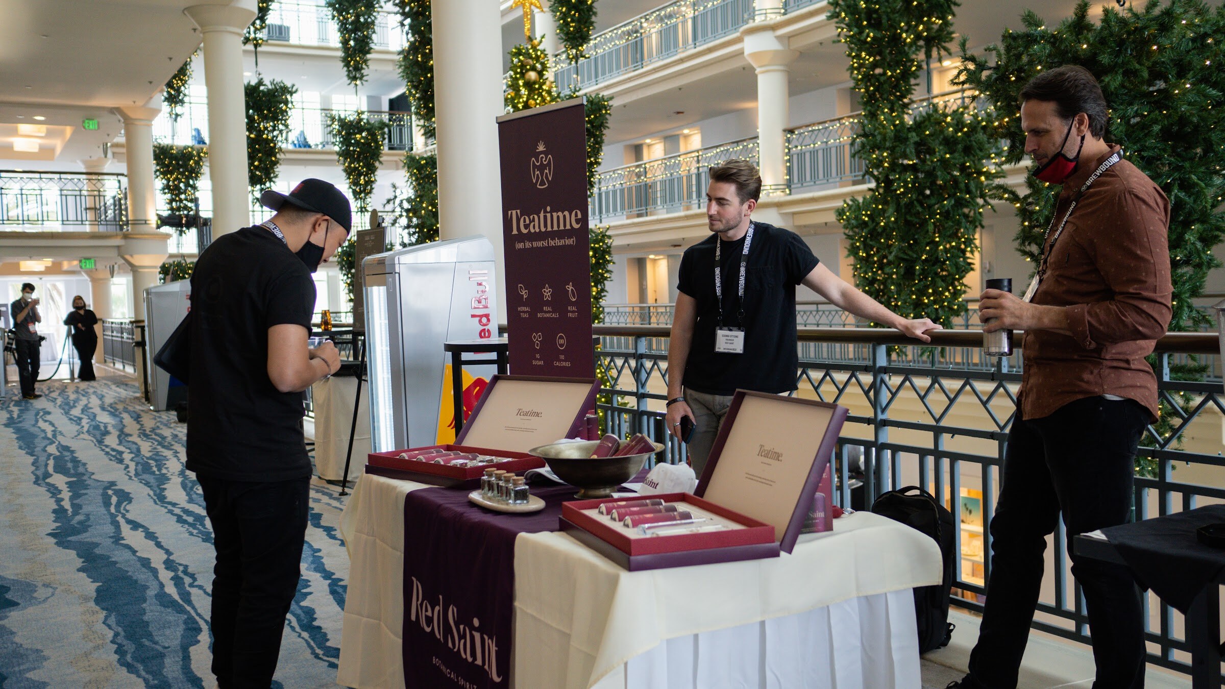 A table with boxed tea samples and a purple Teatime banner on a balcony with garland-wrapped columns and three people standing around it.